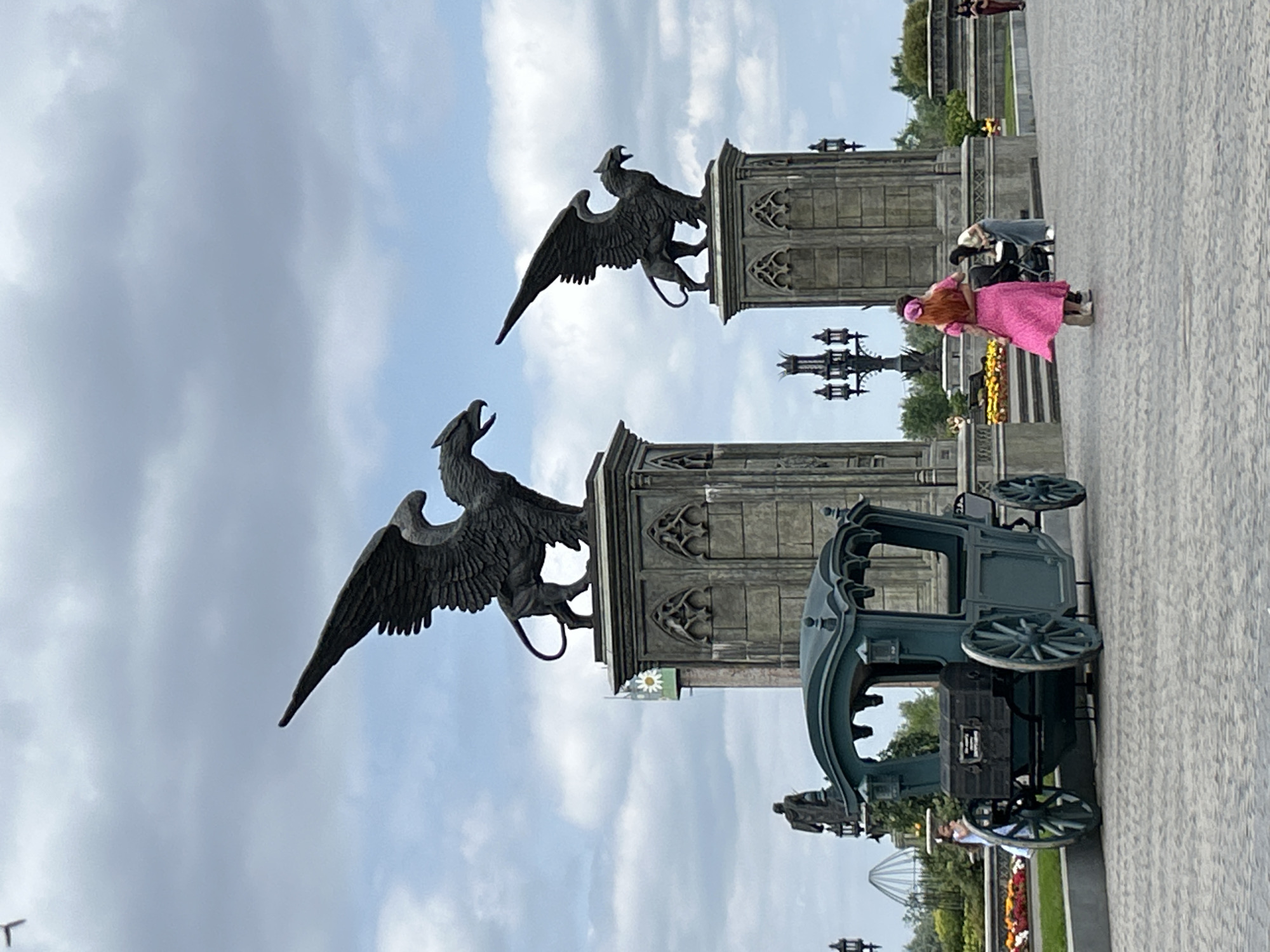 The image depicts a scene in Khryashchevka, Russia, featuring two striking sculptures of griffins perched atop ornate stone structures, likely part of a monument or architectural ensemble. In the foreground, a dark green carriage, possibly a prop or a display, is positioned on a paved walkway. A person dressed in a vibrant pink outfit and a headscarf stands near the carriage, facing away from the camera, with another person partially visible behind them. Further in the background, more stone structures and greenery can be seen, along with hints of a fence and what appear to be amusement park-like structures. The sky is cloudy, suggesting overcast weather. The overall impression is a tourist or public space with a historical or fantastical theme, potentially a theme park or a historical reenactment site. The time of day appears to be daytime, given the ambient light.