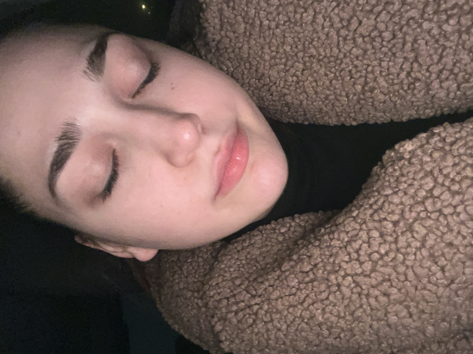 A woman with closed eyes rests her head against a brown, textured, sherpa-like fabric. She has light skin, defined eyebrows, and soft pink lips. She wears a black turtleneck sweater under the fuzzy fabric. The background is dark, highlighting her face and the material she's leaning on. The overall composition suggests a moment of rest or sleep.
