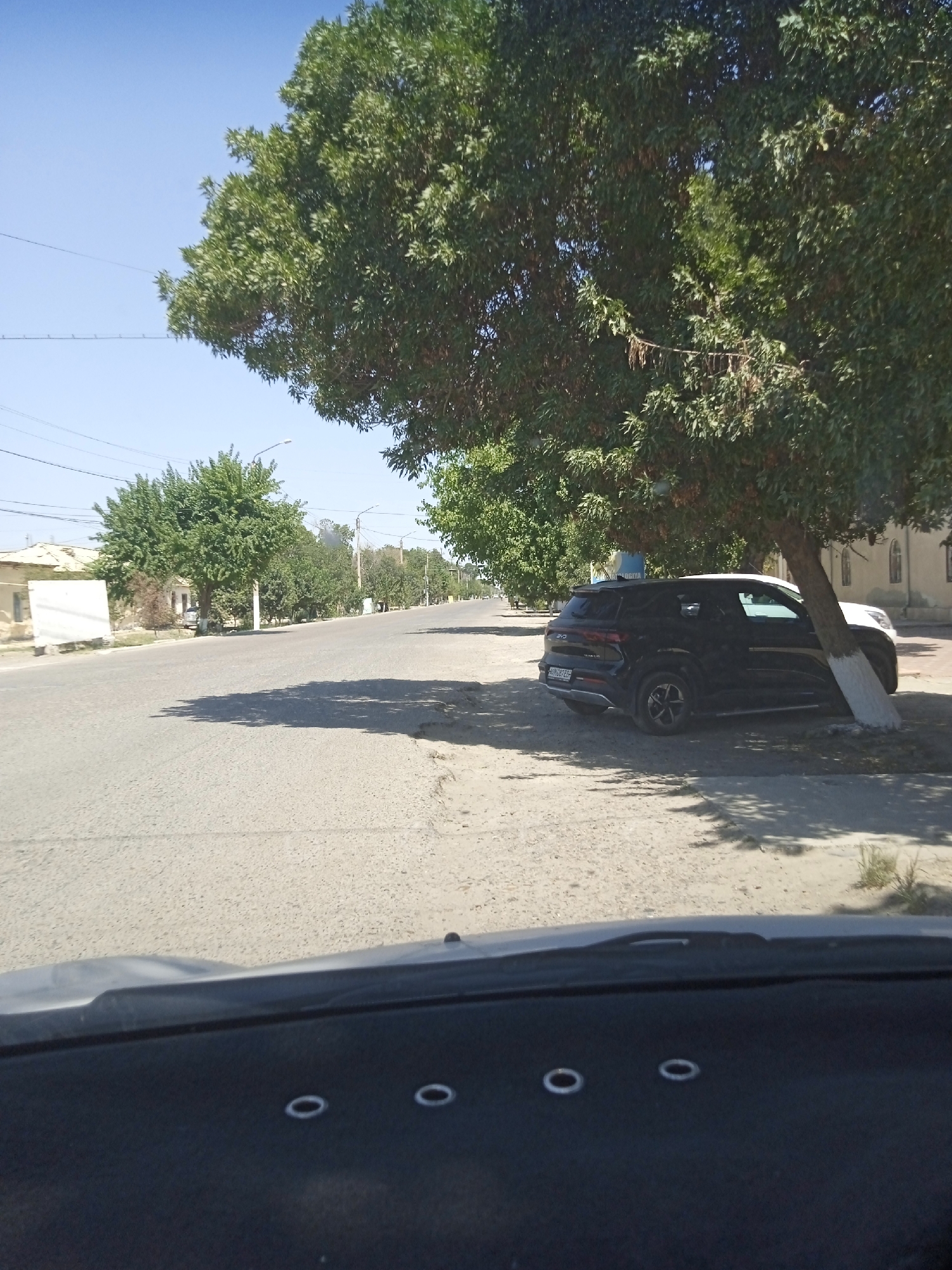 A black SUV is parked under a large tree on a sunny day, casting a shadow on the asphalt road. The car, with license plate "0812 EE", is positioned close to a white building. Trees line the street, and the sky is clear and blue. In the foreground, the dashboard of a vehicle is visible with several circular buttons.
