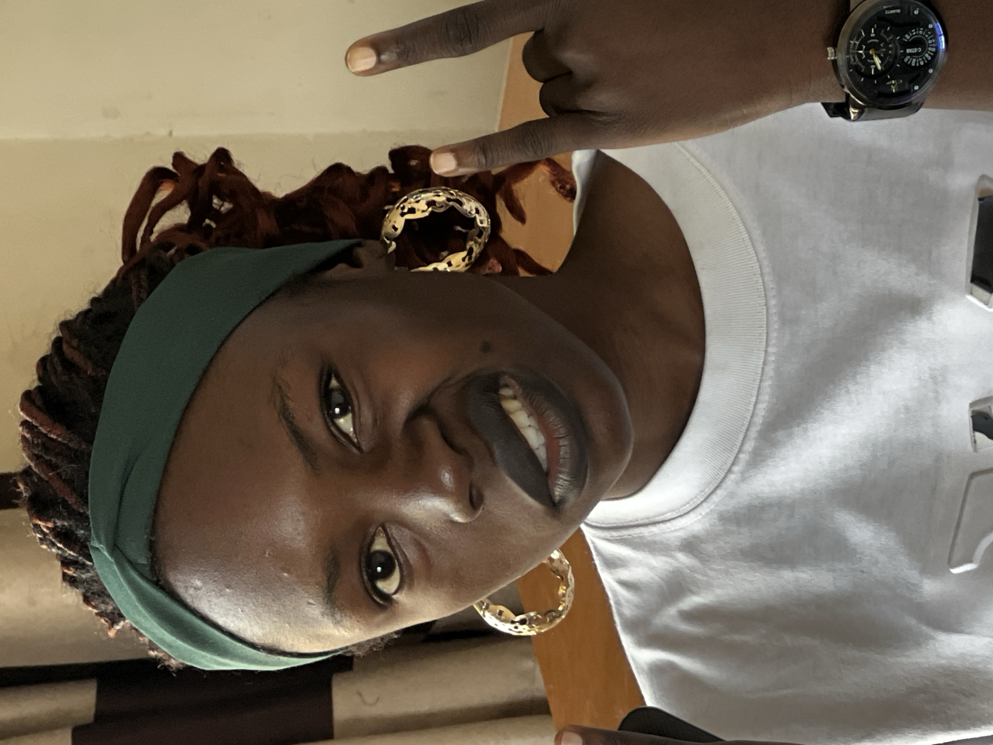 The image shows a close-up, chest-up portrait of a young Black woman wearing a white t-shirt and a green headband. She has her hair in dreadlocks and is wearing large, gold hoop earrings. Her expression is defiant, with her mouth open and teeth bared, and she is making a rock-on hand gesture with her right hand. Her left hand is not visible. The background consists of a plain, light-colored wall and a darker curtain. The lighting suggests an indoor setting, possibly a room in a residential building. The overall impression is one of a candid, informal photograph capturing a moment of bold self-expression. There is no specific visual information in the image that indicates the location within Jos, Nigeria.