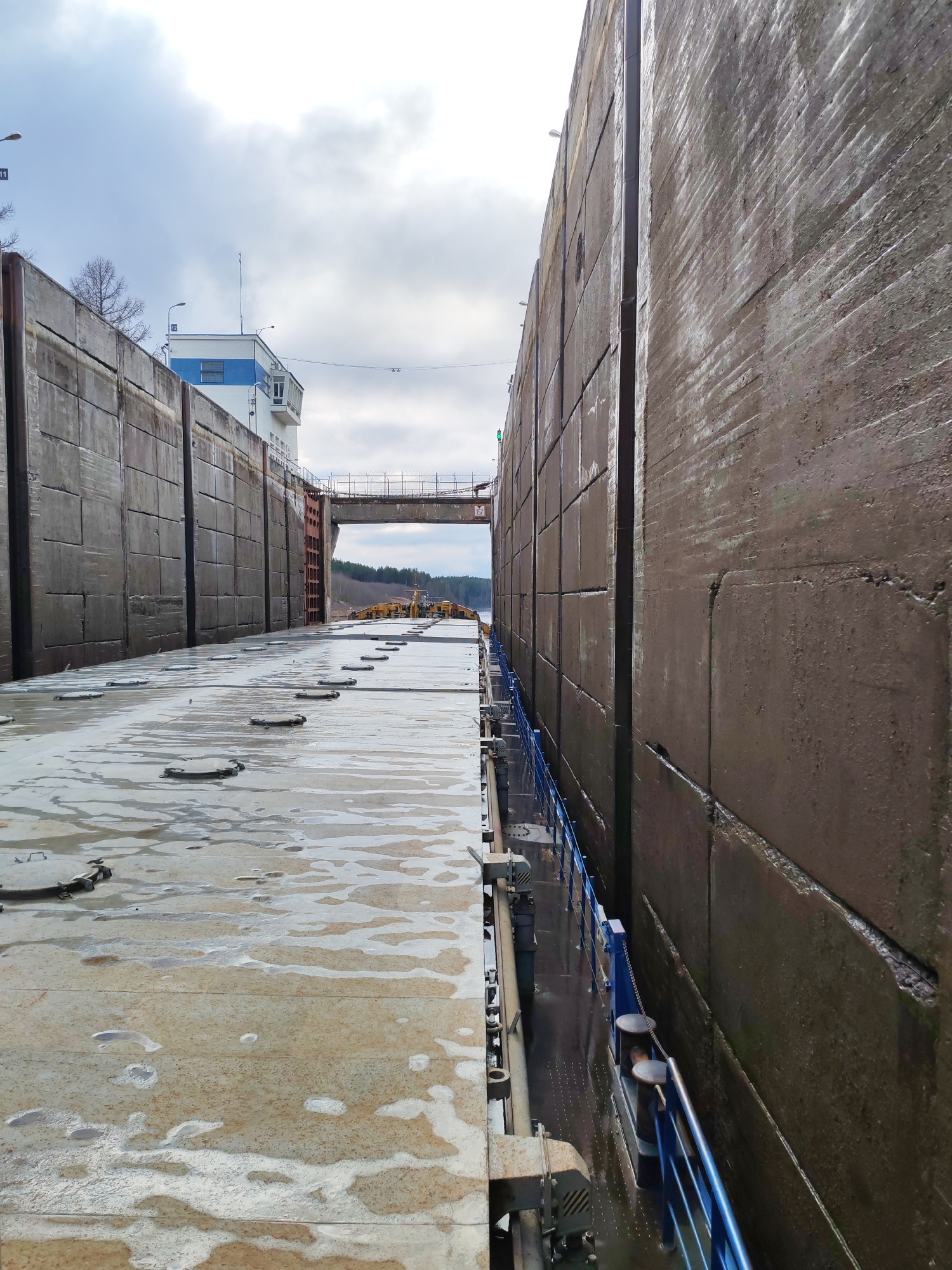 The image depicts the interior of a ship lock, showcasing the large concrete walls that confine the vessel. The floor of the lock is damp and has circular metal access points. A blue railing runs along the side, and mooring bollards are visible. In the background, a control tower is present. The sky is overcast. The location is Unknown, Unknown.
