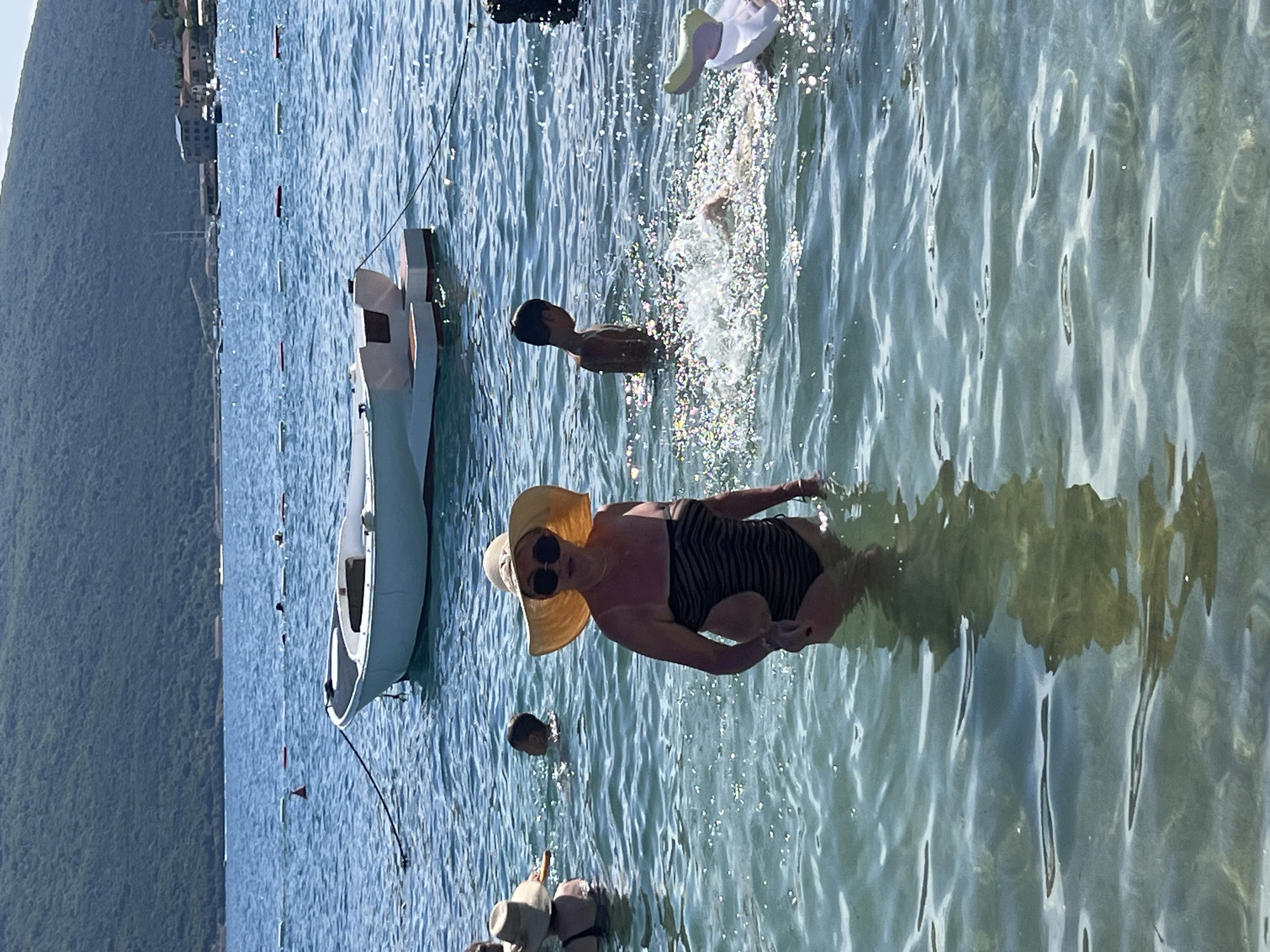 In the city of Đenovići, Montenegro, a group of people are enjoying a sunny day at the beach. The water is clear and shallow, with a gentle ripple. In the foreground, a woman wearing a black and white striped swimsuit and a wide-brimmed straw hat stands in the water, looking at the camera. Her reflection is visible in the water. Behind her, a young boy is splashing in the water, creating a spray of water droplets. Further in the background, a white boat is moored in the water, and a lush green hillside with buildings is visible in the distance. The scene is serene and idyllic, with a sense of peace and relaxation.