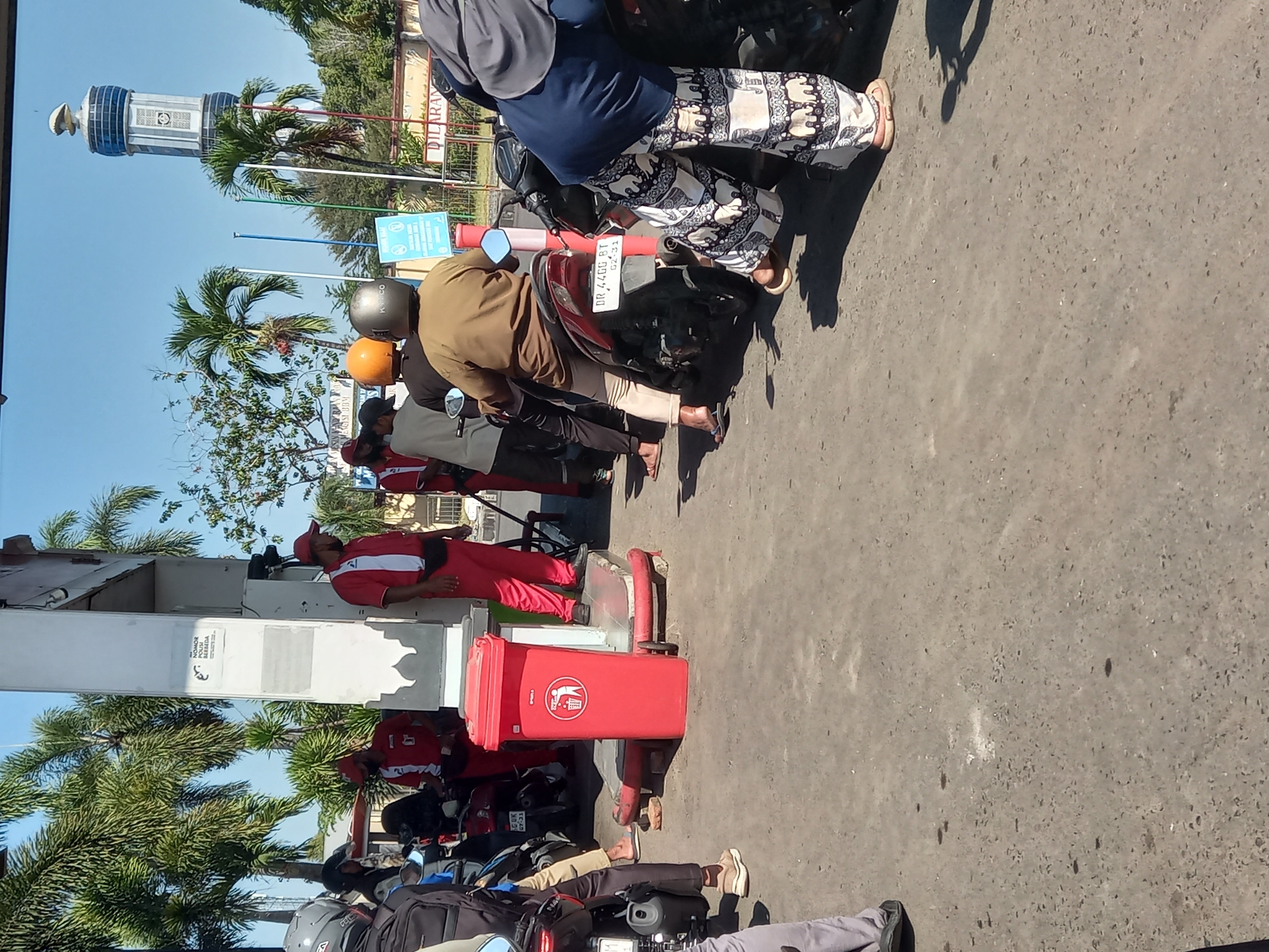 In Mataram, Indonesia, at a gas station, several people are visible. A gas station attendant wearing a red shirt and red pants stands near a fuel pump. Several customers on motorcycles are waiting for service. One customer, wearing a brown shirt and orange helmet, is on a motorcycle. Another customer, wearing patterned pants, is standing next to a motorcycle. A red trash can is also present. In the background, palm trees and a tall, tower-like structure are visible, suggesting an outdoor urban setting. The sky is clear and blue.