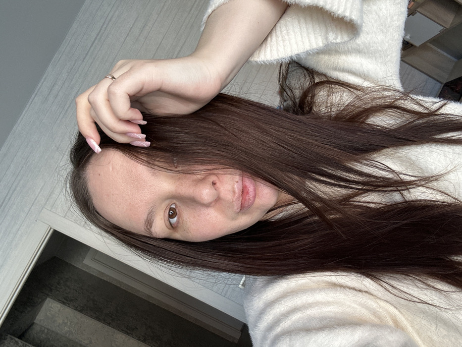 The image displays a close-up, frontal view of a single female subject indoors. The subject has long, dark brown hair, which partially obscures the right side of her face. Her visible left eye is brown. Her skin shows some freckles and minor facial blemishes. Her lips are slightly parted, revealing a visible lesion or cold sore on the upper lip. She wears a light-colored, possibly cream or off-white, fuzzy textured garment, likely a sweater or robe. Her left hand is raised, with fingers near her forehead, appearing to be in the process of adjusting her hair. The nails on her left hand are manicured with an almond shape and a light-colored polish, possibly with a subtle white or pink design. A ring is visible on her left ring finger. The background is blurred but reveals an indoor domestic setting. To the upper left, a light-colored doorframe or wall corner is visible, beyond which a staircase with dark steps and lighter risers is partially discernible. The wall texture in the background suggests wallpaper or a similar finish. The specific location within the city of Chaikovsky, Russia, is not discernible from the image content.
