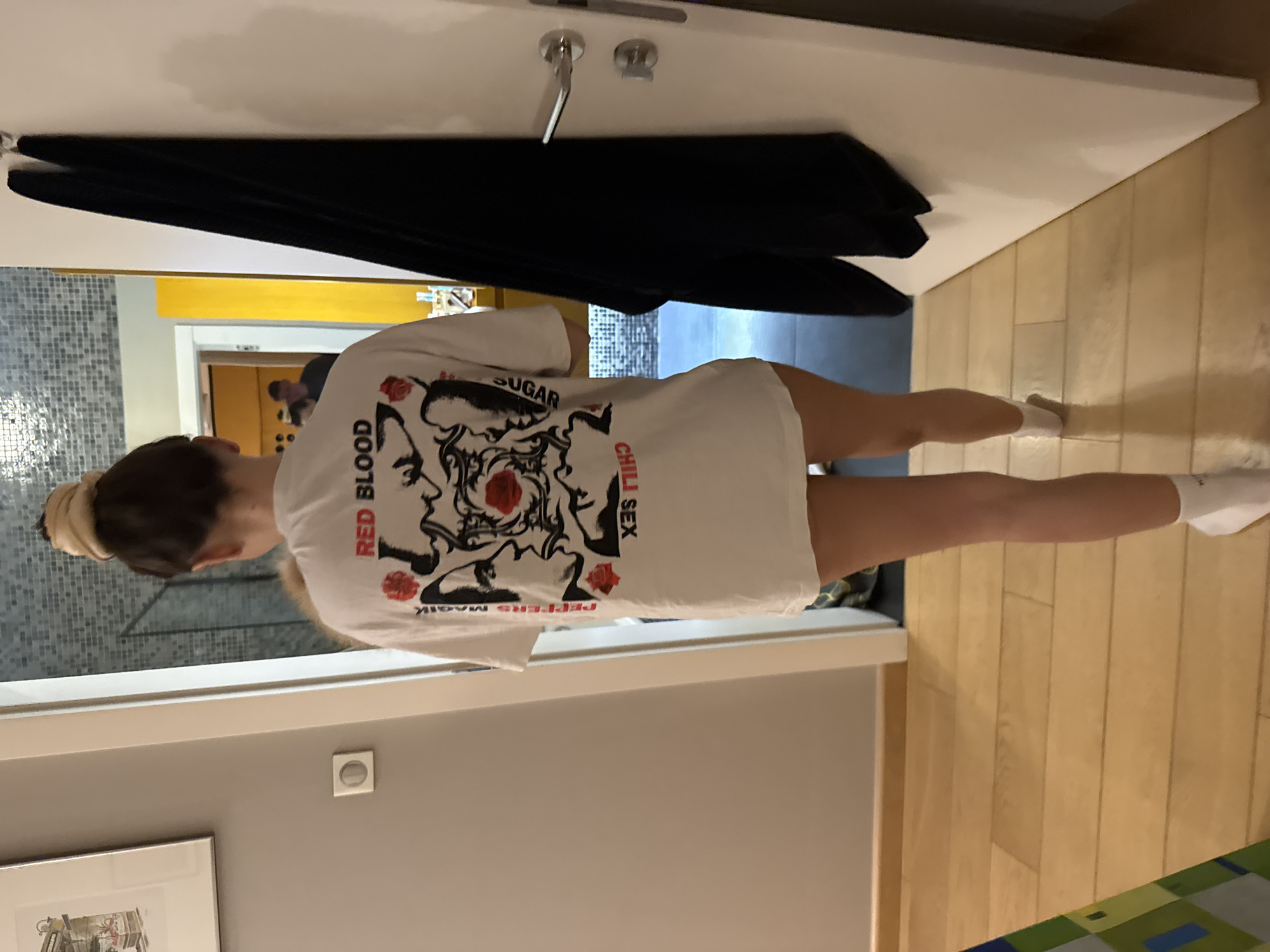 A person stands in a room, wearing a white t-shirt with the words "RED BLOOD SUGAR CHILI SEX" printed on the back. The room has a gray wall, a door with a silver handle, and wooden floors.