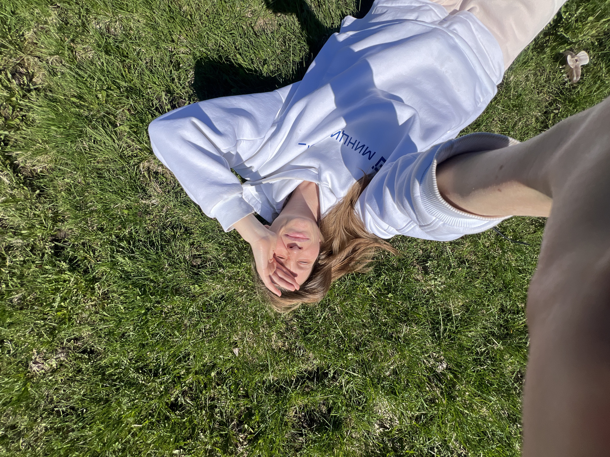 A woman lies on a green lawn, face upward, obscuring her eyes with her right hand. She wears a white hooded sweatshirt and light-colored pants. The grass appears to be well-maintained. The presence of a hand holding a camera suggests a selfie. There is a hair clip on the ground. The image was taken outside in the city of Раздолье, Russia.