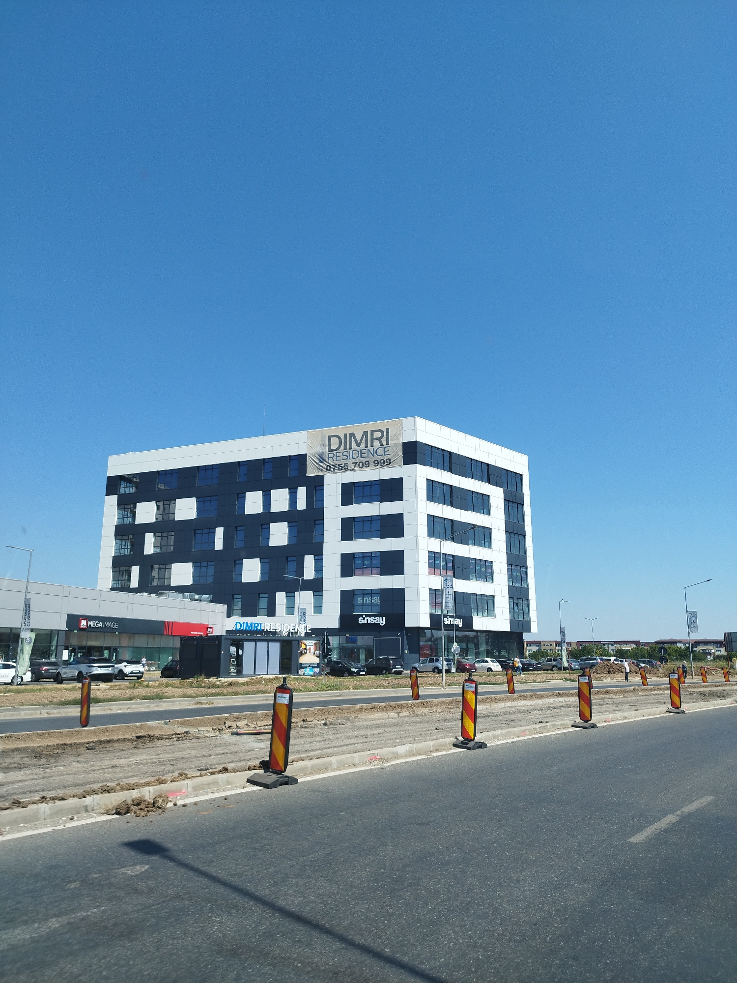 The image shows a modern, multi-story building with a distinctive black and white facade, bearing the "DIMRI RESIDENCE" signage and a phone number. Adjacent to this building is a lower structure with "MEGA IMAGE" prominently displayed. The street in the foreground appears to be undergoing construction, with traffic cones and excavated earth visible. Several cars are parked or driving near the buildings. The sky is clear and blue. The scene is located in Bragadiru, Romania, with the buildings situated on a main road where infrastructure work is underway.