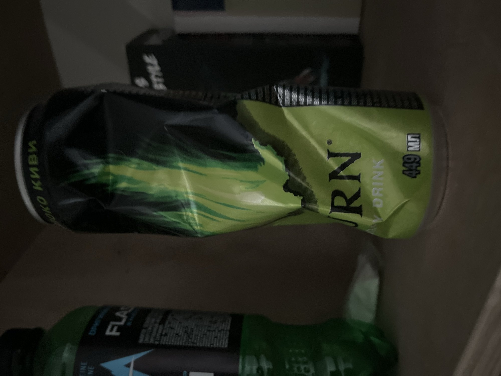 The image shows a partially crushed can of "Burn" energy drink, kiwi flavor, with a volume of 449 ml. It is located next to a bottle of "Flash" energy drink. The drinks are likely placed indoors. The location is unknown, as the image does not provide visual clues about the city of Chaikovsky, Russia. There are no visible people, actions, or interactions.