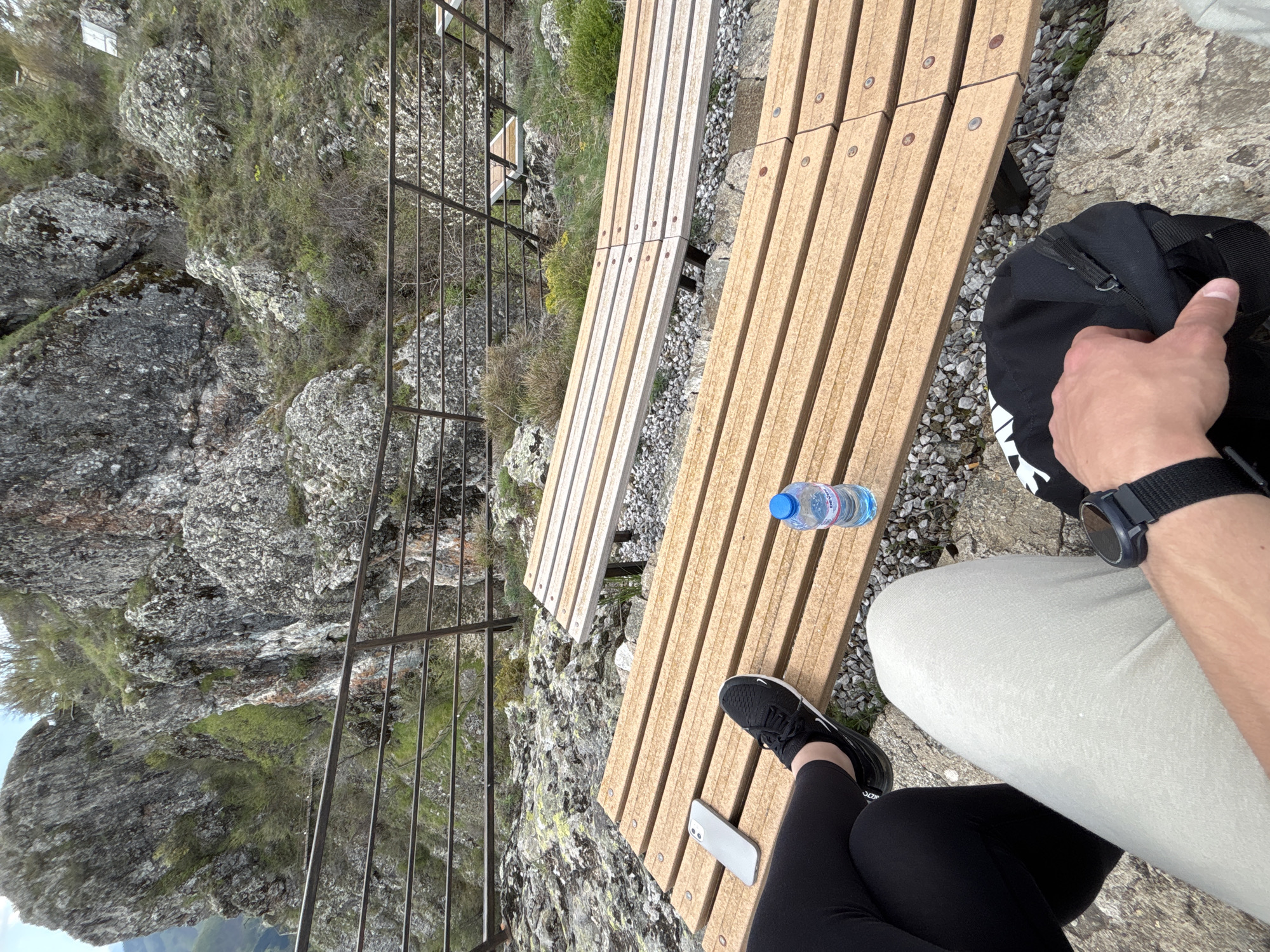 On a rocky mountainside in Gradat, Bulgaria, a wooden bench overlooks a steep drop secured by a metal railing. A person wearing khaki pants and a black watch sits on the bench, their arm resting on a black backpack. Next to them, a second person wearing black leggings and black sneakers sits with a white smartphone on the bench. A bottle of water sits between them. The background features rugged rock formations with sparse vegetation.