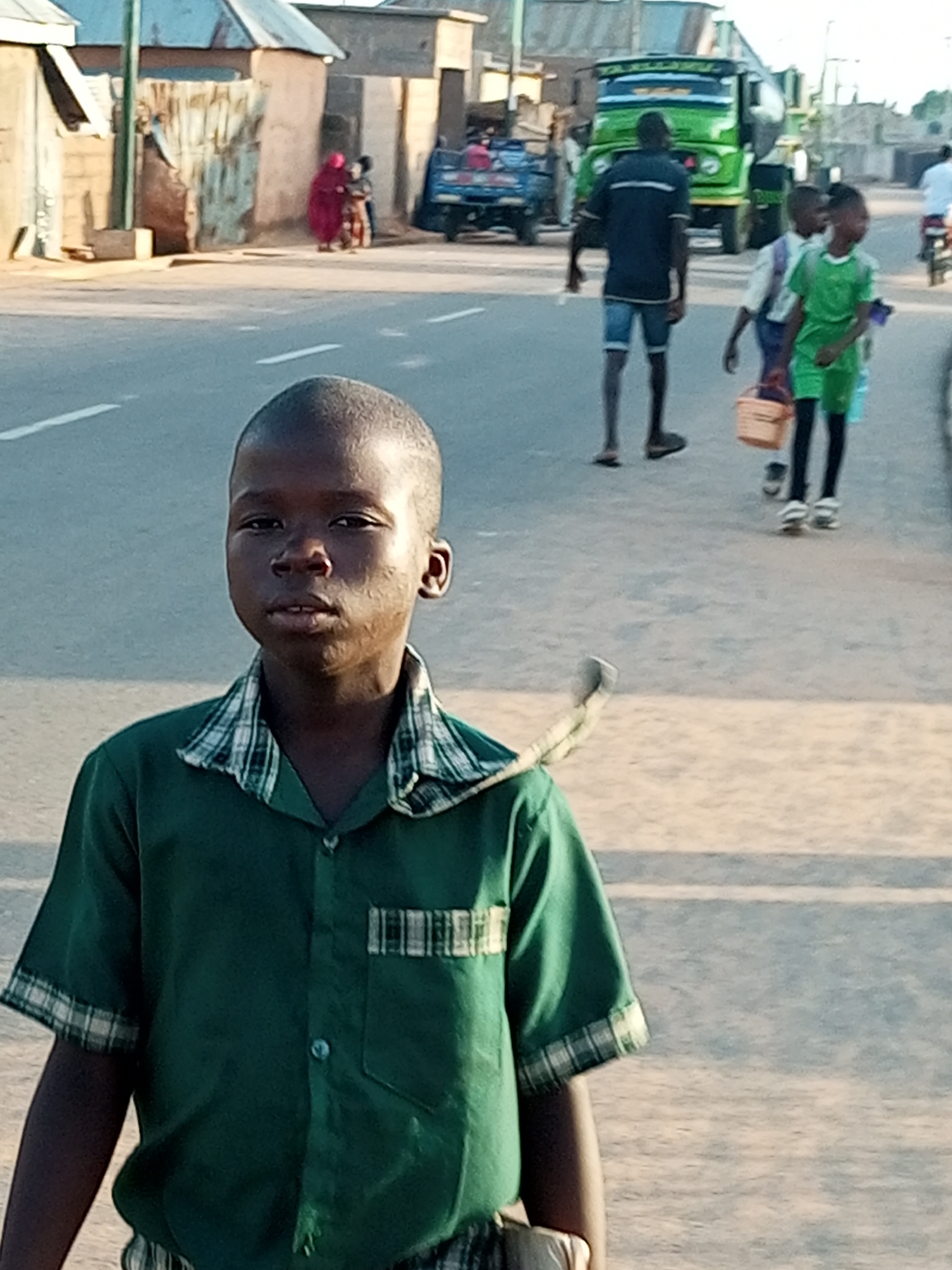 This is a medium shot of a young boy in the foreground, looking directly at the camera. He is wearing a green short-sleeved shirt with a checkered collar and trim. His skin is dark, and he has short hair. The background shows a dusty street scene in the city of Gombe, Nigeria. Several other people are visible, including two young boys walking away from the camera, one carrying a woven basket. A large green truck is parked on the side of the road, and a blue truck is visible in the distance. Buildings with corrugated metal roofs line the street, and some debris is scattered on the ground. The overall lighting suggests it is daytime, likely in the late afternoon due to the warm tones.