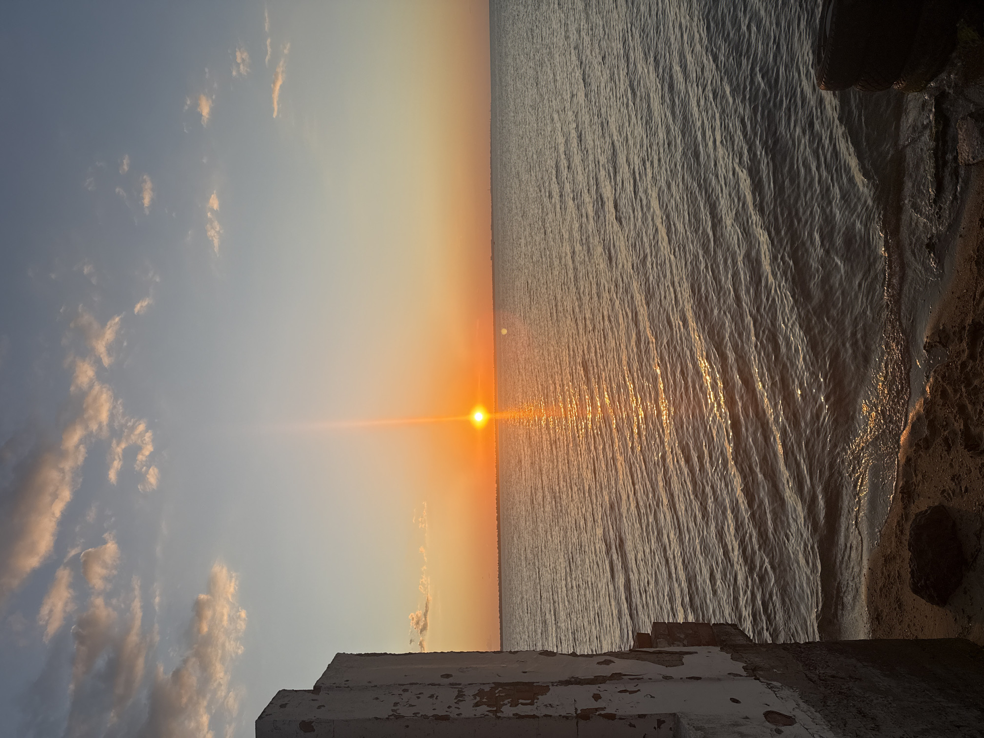 The image captures a sunset over the sea in Zatoka, Ukraine. The sun is low on the horizon, casting a warm orange glow across the sky and reflecting on the rippling water. In the foreground, the weathered concrete structure of a building on the left side of the frame partially obscures the view. The structure appears old and shows signs of decay. A small sandy shore is visible at the bottom, with some dark rocks and debris. The sea stretches out to the horizon, where a faint line of a distant cityscape or shoreline is visible. The sky is a gradient of blue and orange, with scattered clouds tinged with pink. The overall scene evokes a sense of tranquility and the end of the day.