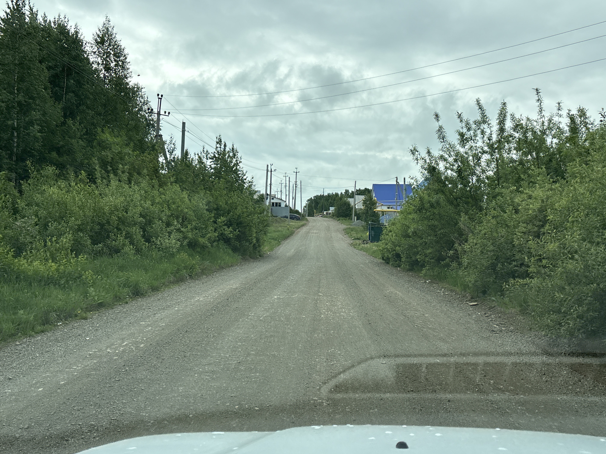A gravel road in Unknown, Unknown leads toward a small village. The road is bordered on both sides by dense green vegetation and telephone poles with wires running overhead. The sky is overcast with a layer of gray clouds. In the distance, a few houses with blue roofs can be seen. The view is from inside a vehicle, looking out through the windshield.