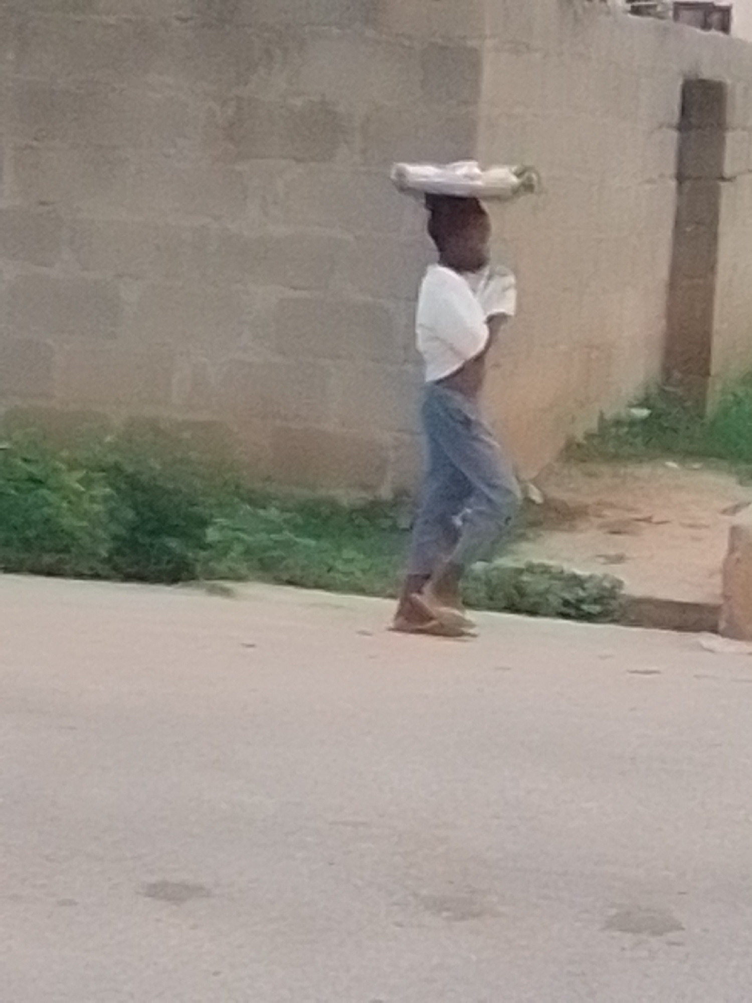 A side profile of a young person is visible, walking on a paved surface. The person is carrying a flat object on their head, possibly food or merchandise, which appears to be covered by a white cloth. They are wearing a white t-shirt and blue trousers that are rolled up at the ankles. The person is wearing open-toed footwear. The background consists of a textured, light-colored wall, with some green vegetation visible on the ground to the right of the wall. The lighting suggests it is daytime, with the sun casting shadows. The scene appears to be outdoors in an urban or semi-urban environment. The location is identified as Liji Hill, Nigeria.