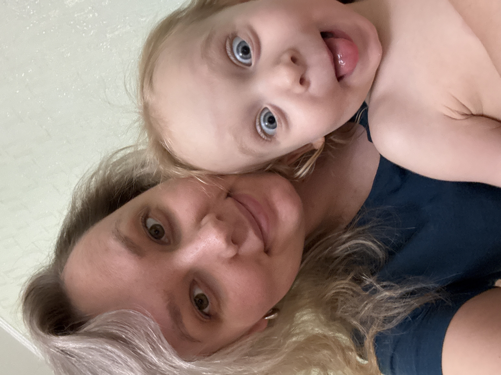 A selfie photograph taken indoors depicts a woman and a young child. The woman is positioned at the bottom of the frame, with her head tilted to the left. Her hair is blonde with graying roots visible. She has light skin and brown eyes and is smiling slightly. The child is positioned above the woman, close to her face. The child has blonde hair and blue eyes and is sticking their tongue out. The child's skin is light. Both are wearing dark blue shirts. The background appears to be a plain, light-colored wall. The image does not provide any specific information to determine a location within Chaikovsky, Russia.
