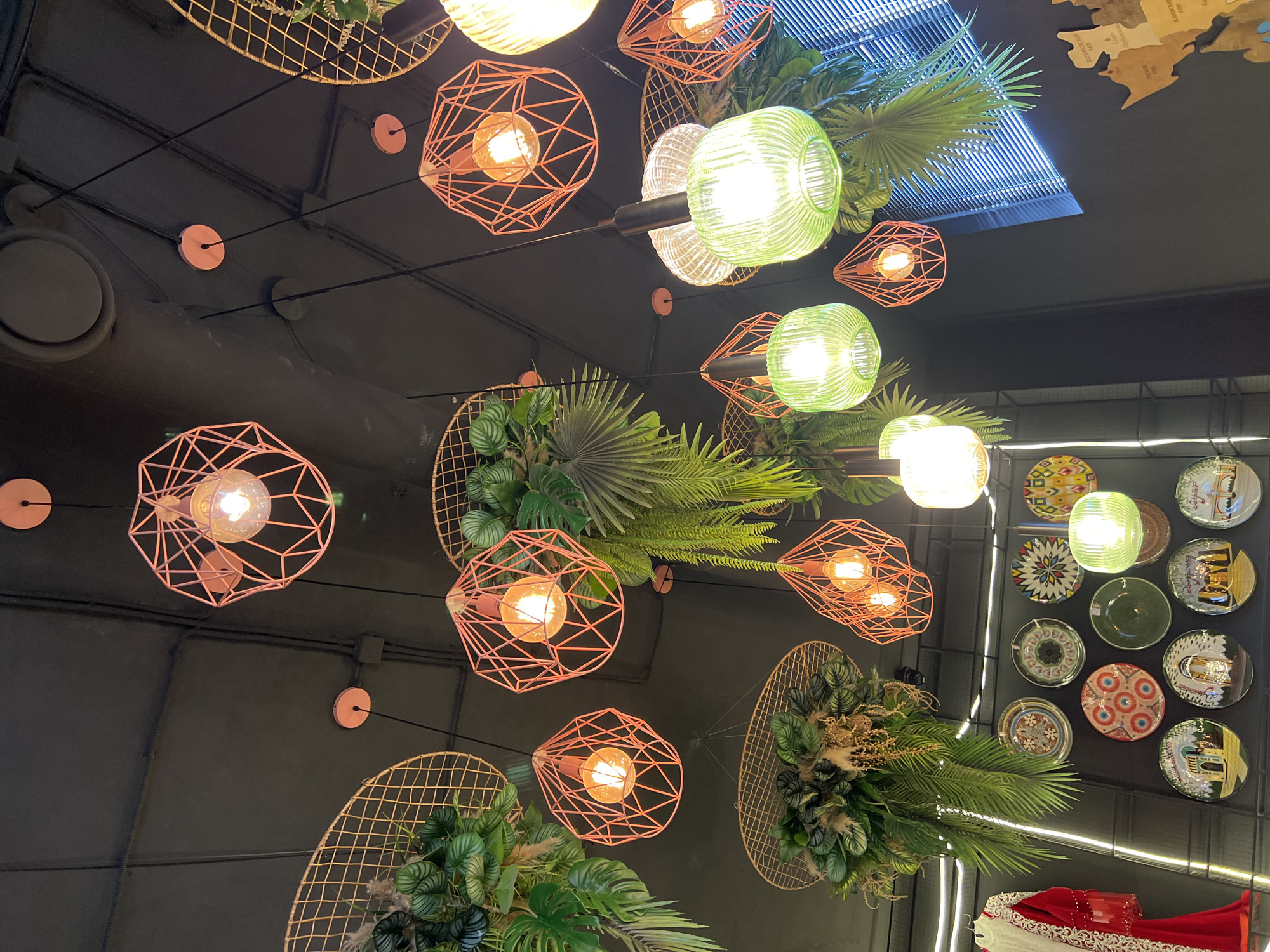 This upward-looking image captures a richly decorated interior ceiling, likely belonging to a cafe, restaurant, or retail space in Krasnodar, Russia. The dominant features are numerous pendant lights, artificial foliage, and decorative elements against a dark gray or black ceiling.
Various light fixtures illuminate the scene. Some are geometric, copper-toned wire cages housing Edison-style bulbs that emit a warm, orange glow. Others are ribbed glass globes, some clear and others with a distinct light green hue, providing a brighter illumination. These lights are interspersed with arrangements of lush artificial greenery, including large palm fronds, ferns, and various broad-leafed plants, often appearing to spill from or be integrated with gold-colored, open-weave decorative elements resembling baskets or screens. Exposed black pipes and small, round, copper-colored mounts for the light wiring are visible on the ceiling.
To the right, a wall is adorned with a collection of decorative plates, each featuring unique and colorful patterns, including abstract designs, floral motifs, and what appear to be architectural sketches. Below these, a vibrant red garment with intricate white and gold embroidered details is partially visible, suggesting a display or a hanging piece of clothing. At the top right, a section of a wooden map with faintly inscribed text is visible, along with a slatted skylight structure allowing ambient light to enter.
No people are present, and no specific activity is taking place, making it a static view of an artfully designed interior. The combination of industrial-style ceiling, warm lighting, and natural-inspired decor creates a lively and inviting atmosphere. No discernible text is clearly readable.