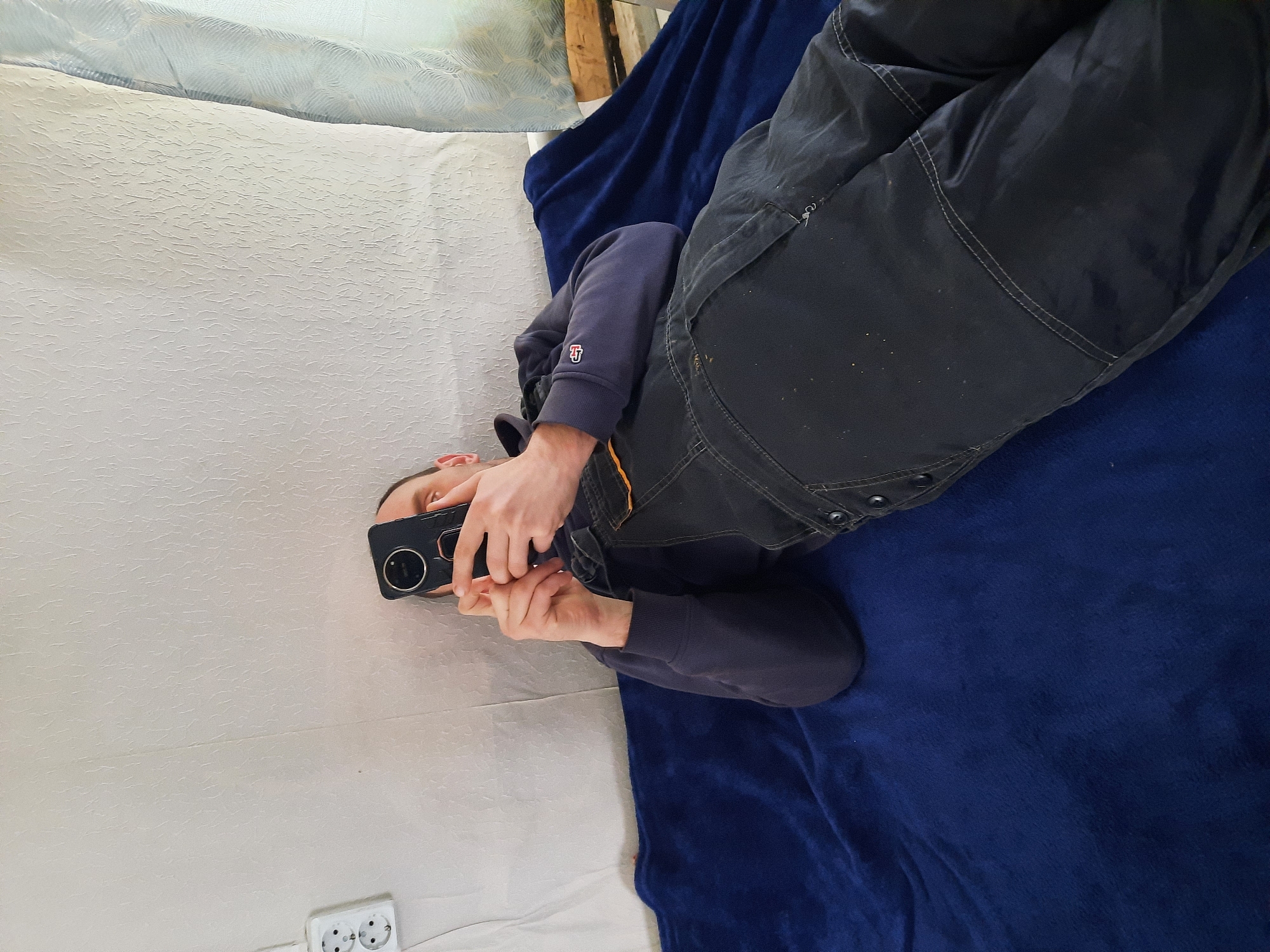 A person lies on a bed, taking a picture with their smartphone. The bed is covered in a white textured blanket and a blue fleece blanket. The person is wearing dark overalls and a dark blue long-sleeved shirt. A white electrical outlet is visible on the wall near the head of the bed. The location is Krasnogorskoye, Russia.
