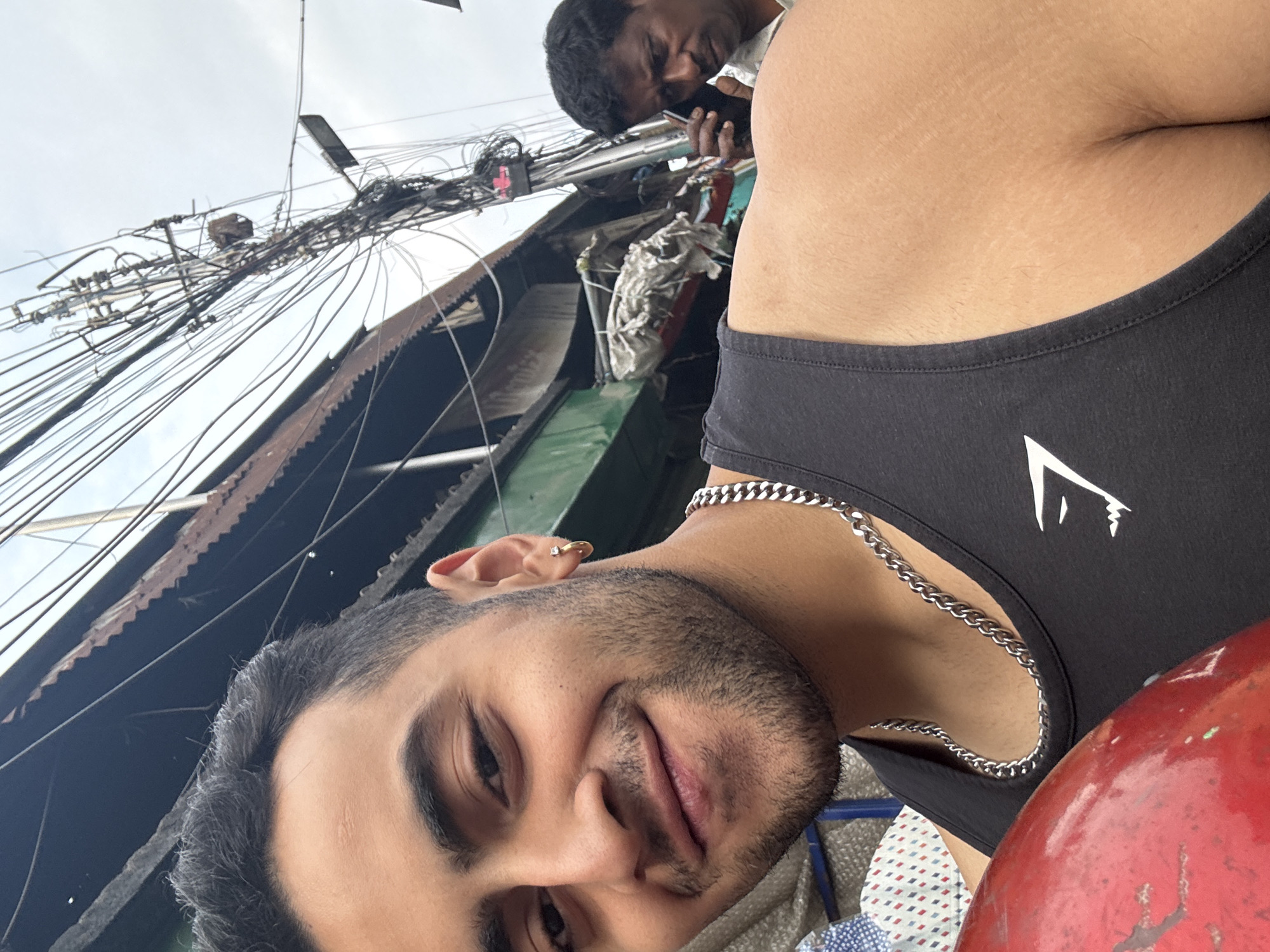 A selfie captures two men against a backdrop of a building with dense, entangled electrical wires. The primary subject is a younger man in the foreground wearing a black tank top with a silver chain. He has a beard and is smiling. A second man, partially visible, is in the background. He is dark-haired and appears to be using a mobile phone. The background building has a corrugated metal roof and a green wall section. The dense network of electrical wires dominates the upper part of the frame. The scene is plausibly in Kolkata, India, based on the style of building construction and the disorganized electrical wiring.