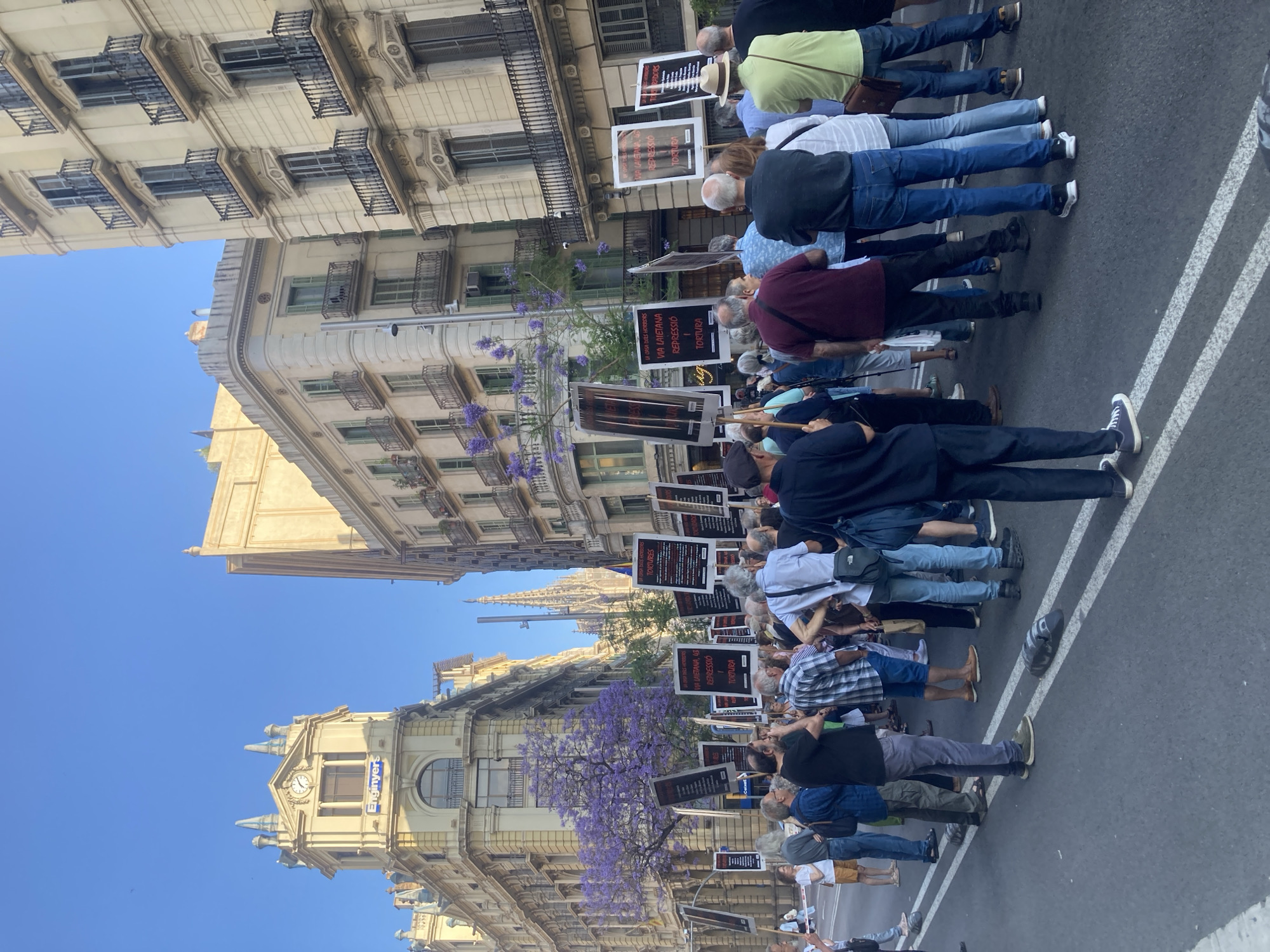 The image depicts a daytime protest or demonstration taking place on a street in Barcelona, Spain. A group of people, mostly adults, are seen from behind, standing in a line along the roadside, facing away from the camera. Many are holding protest signs or placards.
The signs prominently display text in Catalan or Spanish, including "VIA LAIETANA, 43," "REPRESSIÓ" (Repression), "TORTURA" (Torture), "LA CASA DELS HORRORS" (The House of Horrors), and "TORTURES." This indicates the protest is against repression and torture, likely referencing historical events or ongoing concerns associated with the address Via Laietana 43.
The scene is set on an urban street flanked by multi-story buildings with classical architecture, decorative balconies, and shuttered windows. One building features a clock tower and a visible sign that reads "Enginyers." A distant church spire can be seen peeking above the buildings. Vibrant purple flowering trees, likely Jacarandas, are scattered along the street, adding color to the otherwise stone-colored buildings. The sky is clear and blue, suggesting fair weather. White lines marking a lane are visible on the asphalt in the foreground. The overall impression is a collective public action in a historic city environment.