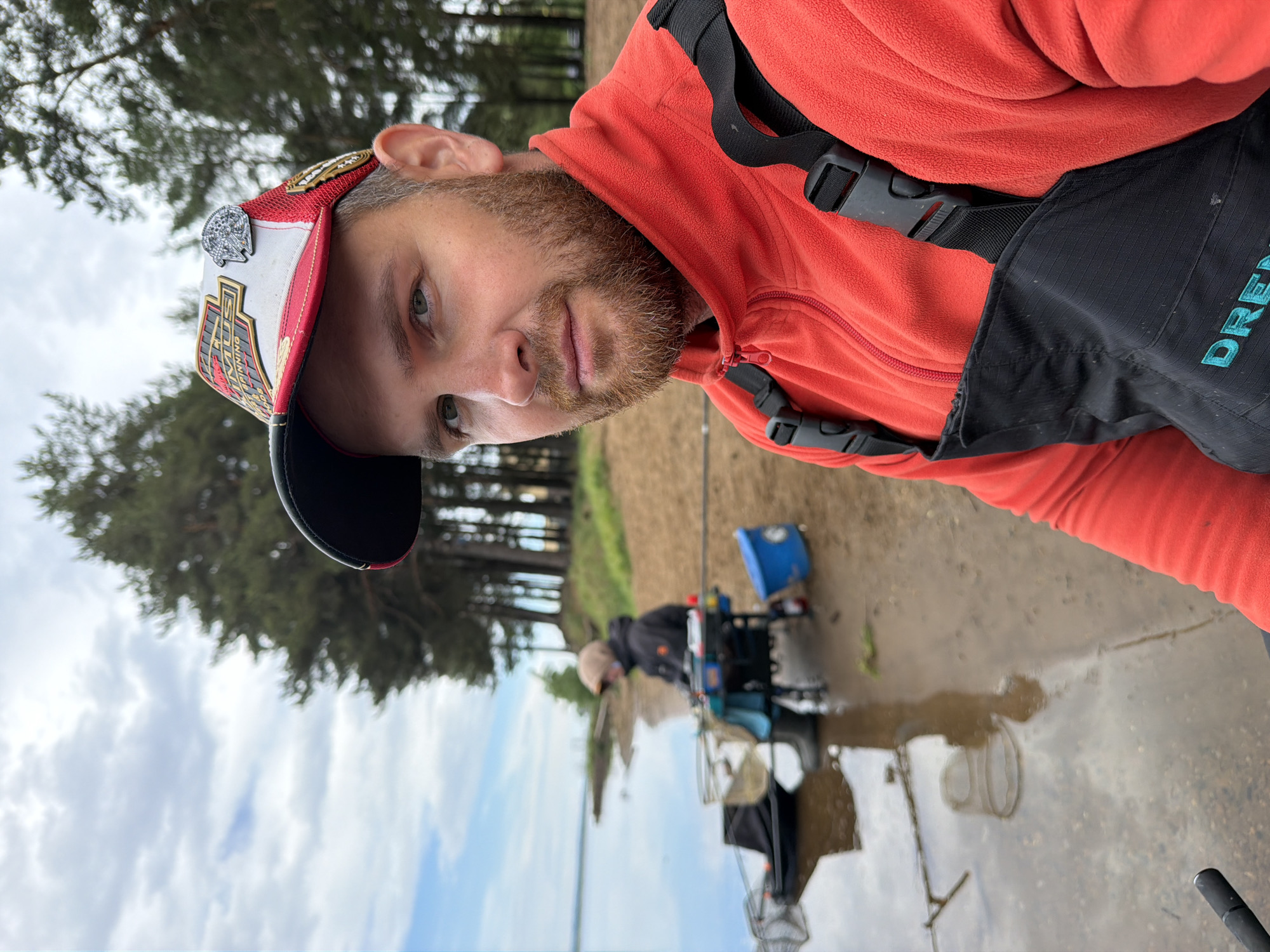 A medium shot captures a man wearing an orange fleece and a red, white, and black baseball cap, likely at a fishing location in Chaikovsky, Russia. The man is facing the camera. Straps are on his chest. Another man is visible in the background, seated in a fishing chair next to the water. Trees are on the bank on the left side of the image. The weather looks overcast and the location appears to be either next to a river or a lake. The foreground is damp.