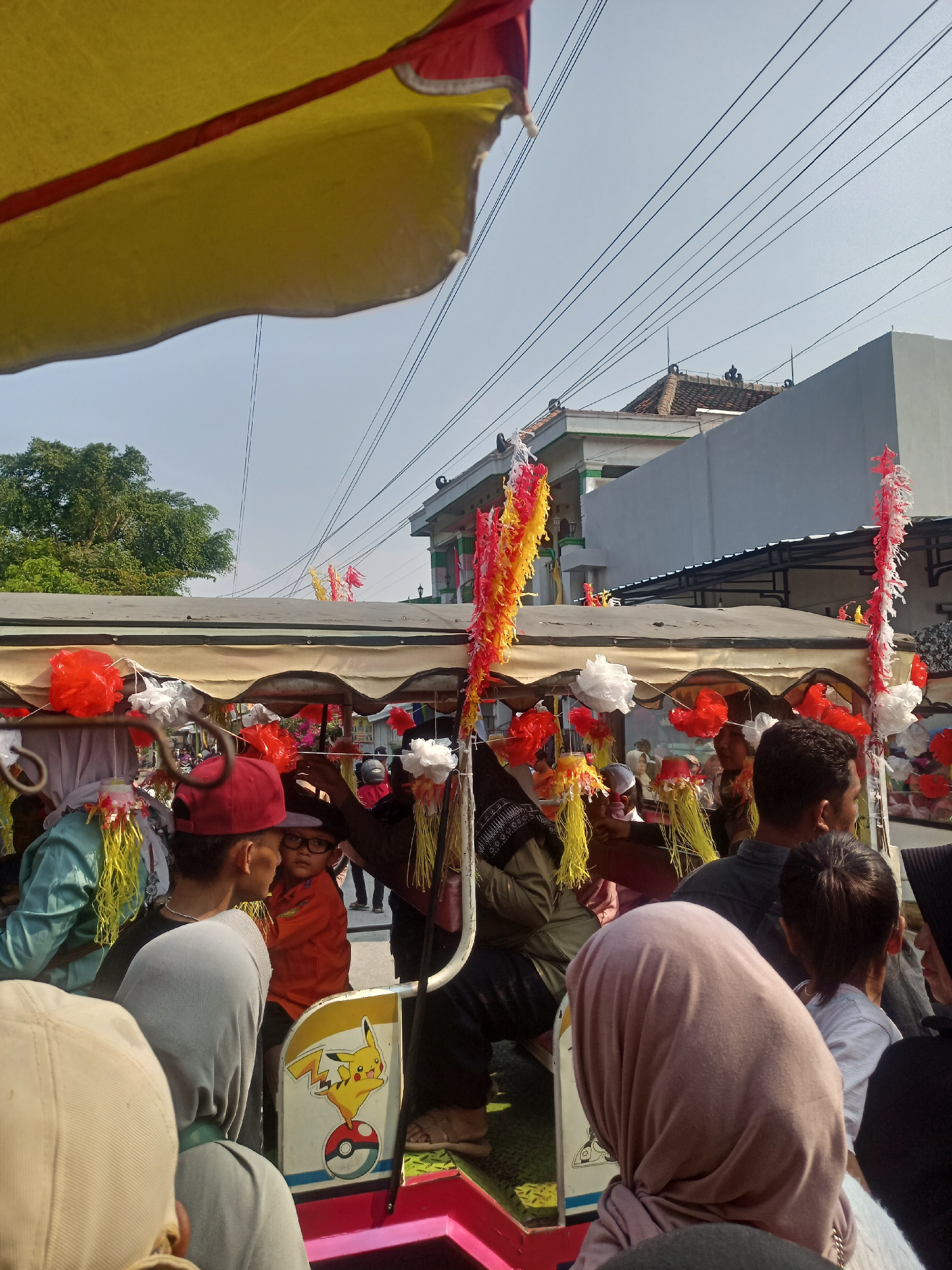 In Gemolong, Indonesia, on a street lined with utility wires and buildings, a festive parade is underway. The foreground is dominated by a colorful, decorated cart, possibly a small truck or rickshaw, adorned with red and white streamers and yellow and red paper decorations. Several individuals are visible within and around the cart, suggesting participation in a procession.
In the cart, a young boy wearing an orange shirt and glasses is seated, looking towards the left. Beside him, an adult wearing a red cap is also visible. Other people are clustered around the cart, some facing away from the camera, indicating they are part of the parade. On the side of the cart, a decal featuring the character Pikachu from Pokémon is prominently displayed.
The background shows a building with a green roof and white facade, typical of Indonesian architecture. Trees are visible on the left, and the sky is clear and bright, indicating a daytime scene. The overall atmosphere appears to be one of celebration and community gathering.