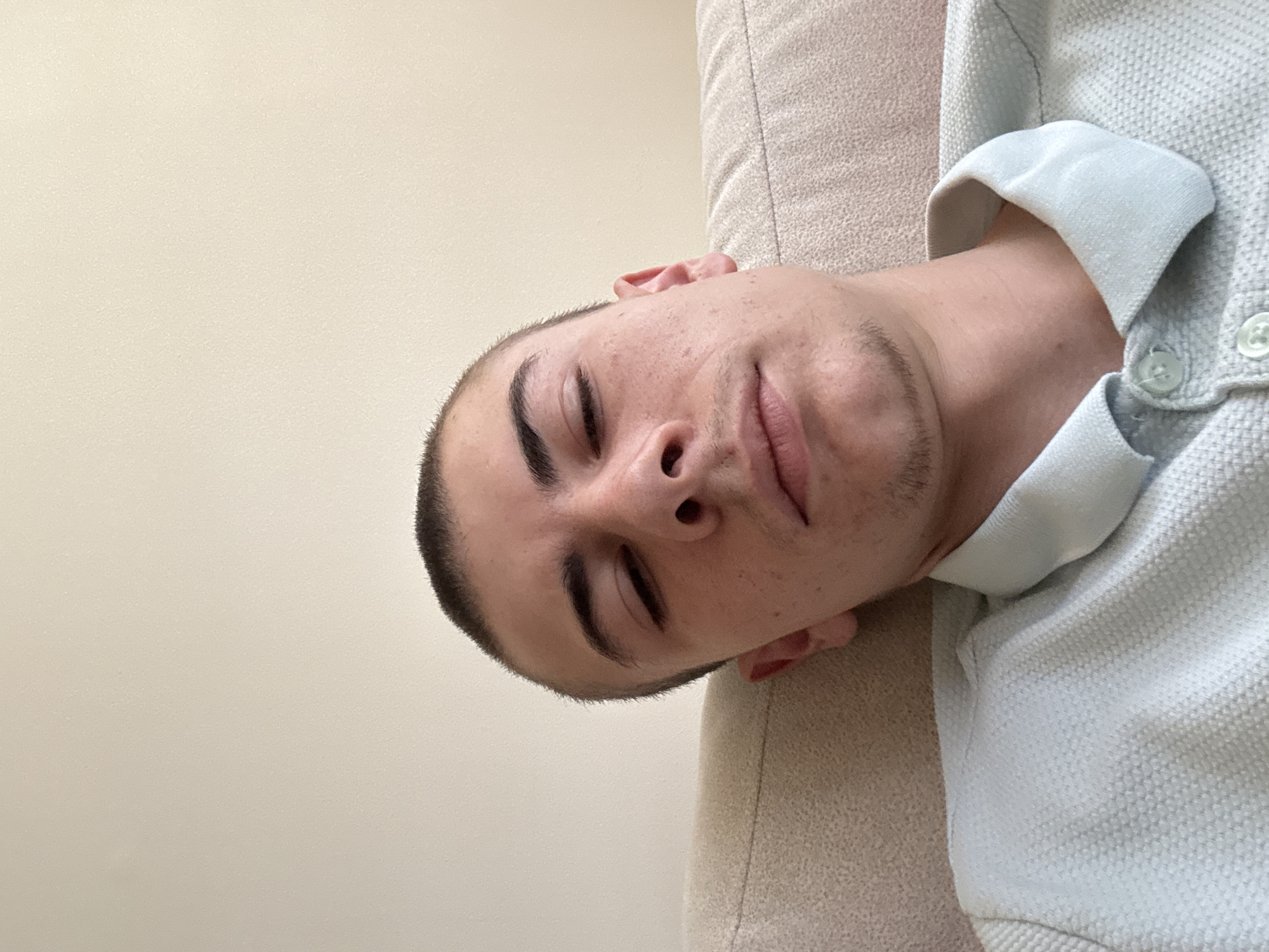 A young man with short, dark hair is lying on a light beige couch. His eyes are closed. He is wearing a light blue collared shirt. The background is a plain, light beige wall. No information about Plovdiv, Bulgaria is discernible.