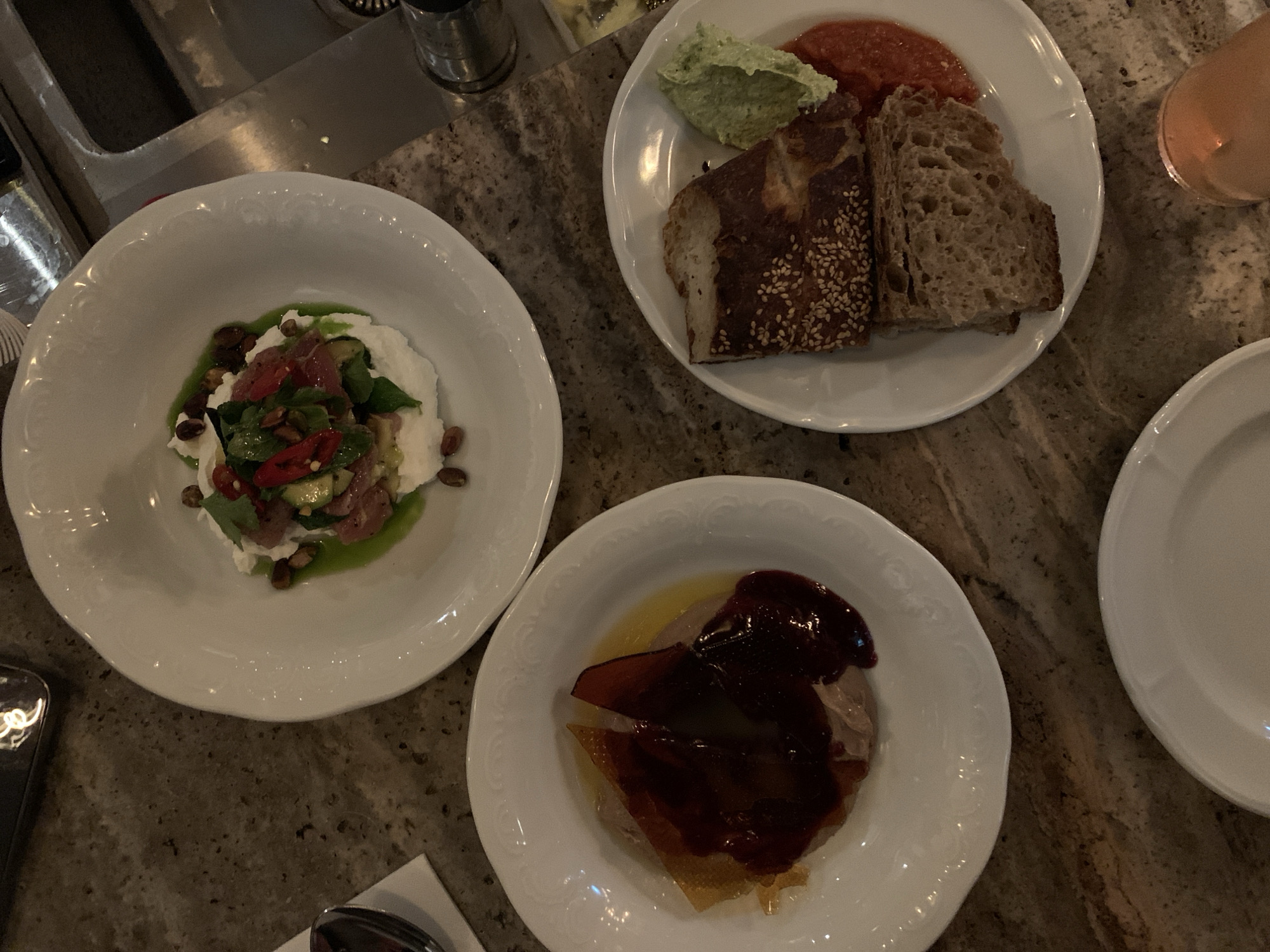 The image shows a close-up, overhead shot of a tabletop scene, likely within a restaurant setting. The table surface is patterned with a speckled brown and gray design, suggesting a stone or composite material. Several plates are arranged on the table, each containing different food items.
- The plate on the left holds a white, creamy substance topped with a colorful mix of greens, reds, and other ingredients, potentially a burrata salad.
- The plate at the top contains slices of bread, a green spread, and a red sauce, potentially hummus, pesto, and tomato chutney.
- The plate at the bottom features what appears to be a glazed duck or meat dish with a dark, rich sauce.
- A white plate can be seen on the right side.
There are also hints of cutlery and beverage in the scene.
The location can be inferred to be in a restaurant in Tel Aviv, Israel.