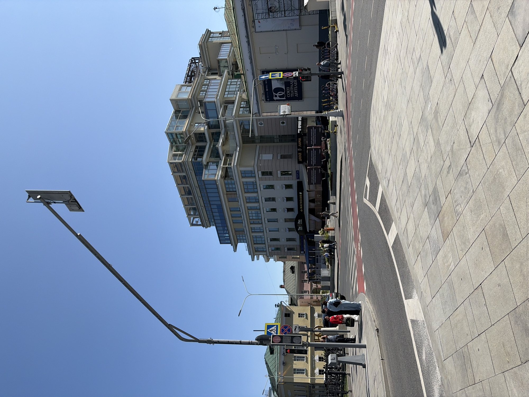This is a street-level, eye-level, daytime, full-shot photograph taken in Moscow, Russia, on a clear day with a bright blue sky. The scene is an urban intersection with a multi-story modern building prominently featured on the right, characterized by its light-colored facade and numerous glass-enclosed balconies. To the left, older, lower buildings with green roofs are visible.
In the foreground, a paved pedestrian area transitions to a road with a red-striped crosswalk. A traffic light pole with pedestrian crossing signals and a bicycle symbol is situated on the sidewalk to the left. Several people are standing near the traffic light pole, including a woman in a red top and a person in a light blue tracksuit. Further down the sidewalk, a group of people are seated on a bench.
The midground shows the crosswalk and the street, with several parked bicycles and yellow electric scooters. The modern building has a sign that reads "The Pho" and another that appears to be in Cyrillic. A large banner on a building to the right of the modern structure displays Cyrillic text and a graphic with the number "150" and "СОЮЗ ТЕАТРАЛЬНЫХ ДЕЯТЕЛЕЙ" (Union of Theater Workers).
The background consists of more buildings and a clear sky. The overall atmosphere is bright and sunny, typical of a pleasant day in the city.