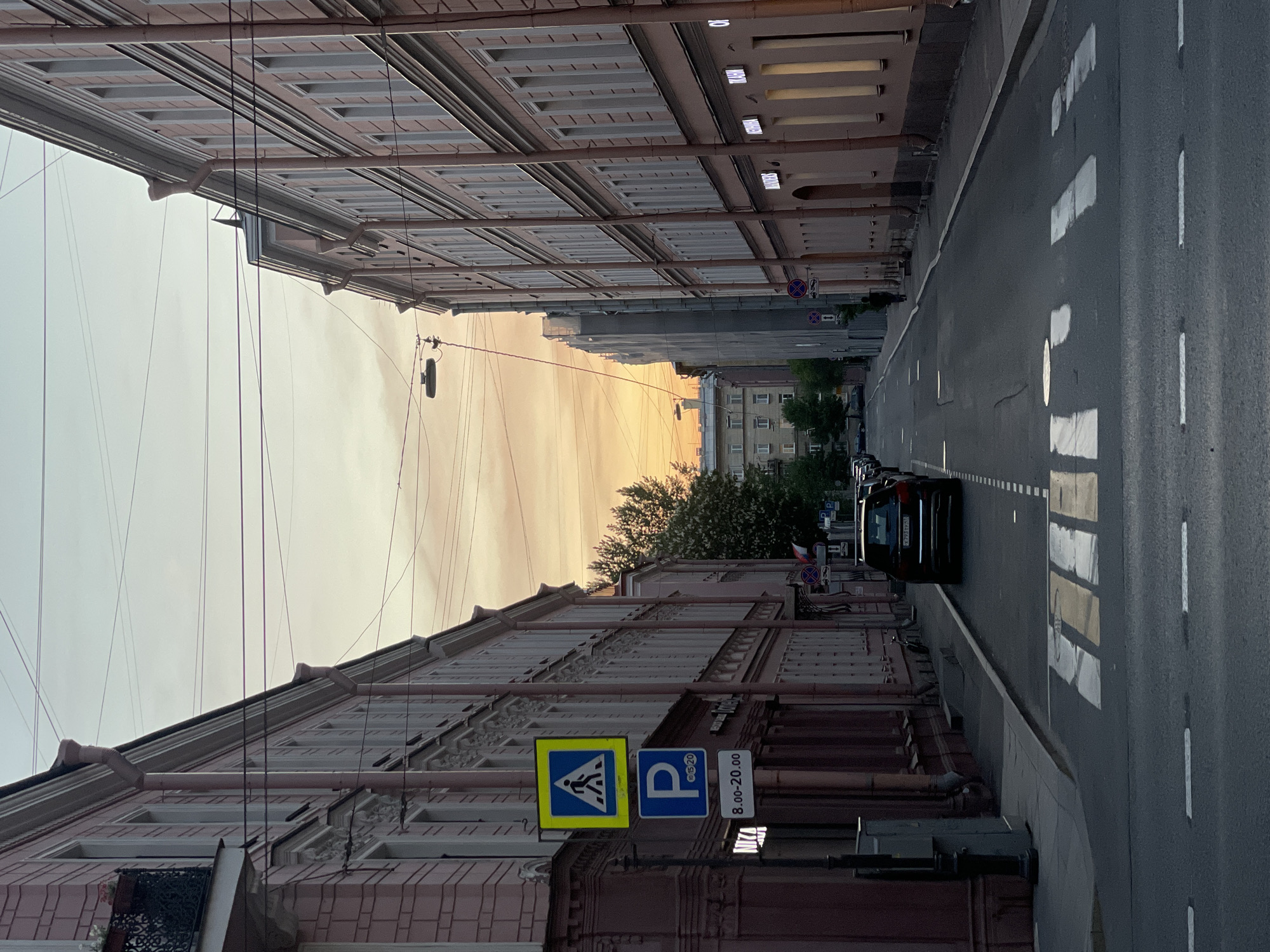 The image shows a street scene in Saint Petersburg, Russia, facing west with buildings lining both sides. The road has crosswalk markings and a black car is driving away in the right lane. A pedestrian crossing sign and parking sign are visible on the lower-right side of the image. The sky is pale with orange hues, possibly indicating sunset. Overhead wires run across the street.