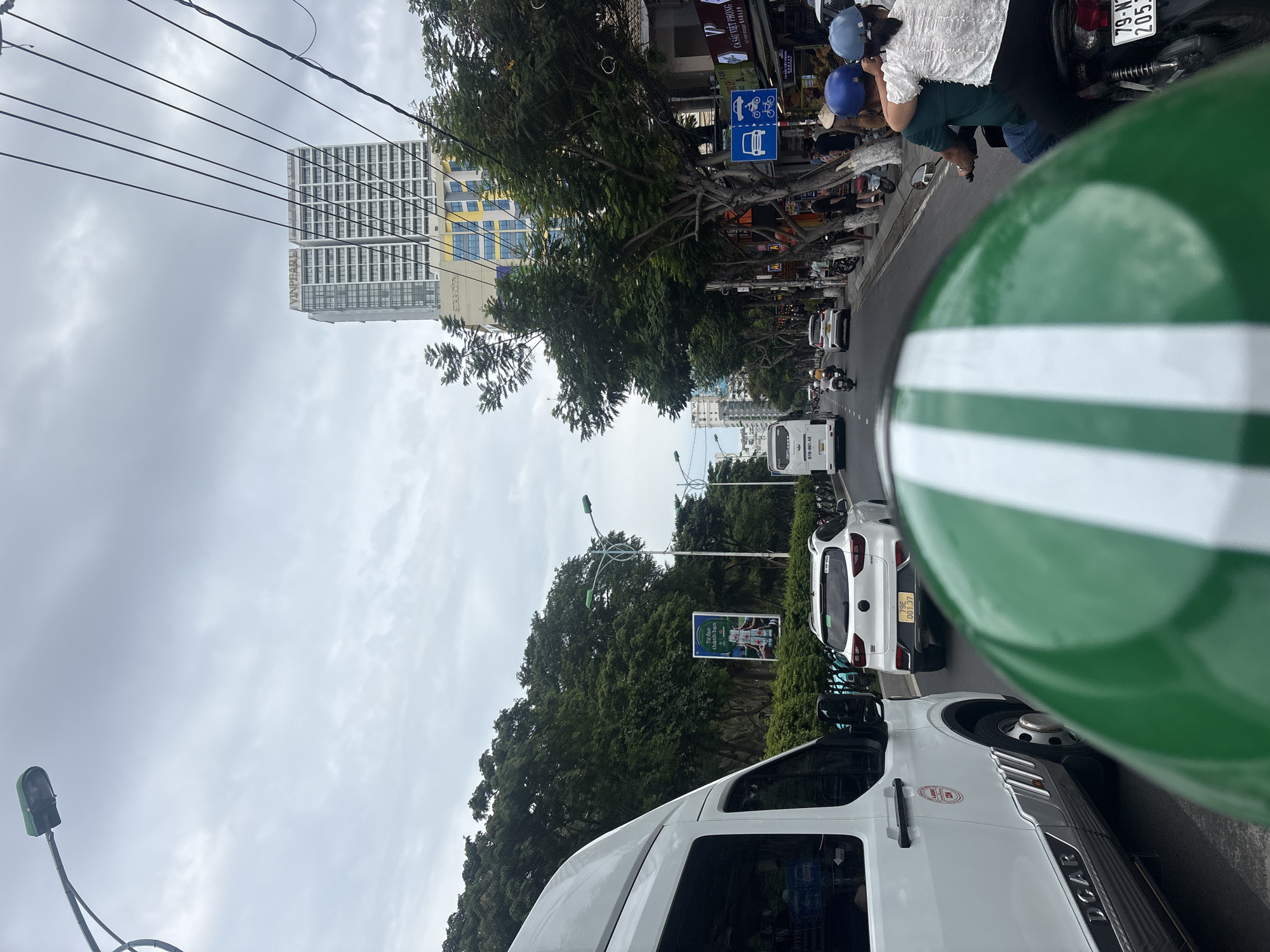 The image depicts a street scene in Nha Trang, Vietnam, characterized by overcast skies and moderate traffic. The perspective is from the back of a motorcycle, as indicated by the green and white striped helmet in the foreground.
In the mid-ground, a white van with "DCAR" branding is prominently visible on the left. To its right, a white SUV is traveling in the same direction. Further down the street, a white bus is also present. A line of trees with dense foliage borders the right side of the road, with several buildings visible behind them. One notable building is a tall, modern high-rise with "VINPEARL" signage.
Several individuals are on motorcycles. In the immediate foreground, a couple is riding a motorcycle, with the person in the back wearing a blue helmet and the person in front wearing a light blue helmet and a white shirt. Other motorcyclists are scattered throughout the scene, some carrying passengers.
Streetlights and power lines are visible overhead. A large billboard advertising an energy drink is positioned between trees on the right side of the road. Traffic signs, including one indicating shared lanes for cars, motorcycles, and bicycles, are also present. The road surface is asphalt. The overall atmosphere suggests a typical urban environment during daytime.