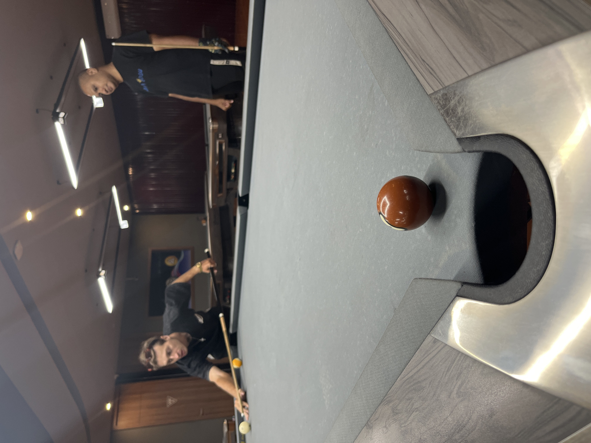 Two men are playing pool in what appears to be a dimly lit bar or billiard hall, suggested by the overhead fluorescent lighting and dark wooden interior. The scene is captured from a low angle, looking across the pool table. One man, with a bald head, is standing and holding a cue stick, seemingly about to take a shot. He is wearing a black t-shirt. Another man, with darker hair and sunglasses, is in the foreground, leaning over the table in a shooting stance, also holding a cue stick. He is wearing a black t-shirt and appears focused on his shot. A single brown billiard ball is visible on the table, positioned near a pocket. The pool table has a grey felt surface and a metallic bumper. The overall atmosphere suggests a casual game of billiards. The image is oriented vertically. There is no visible text in the image.