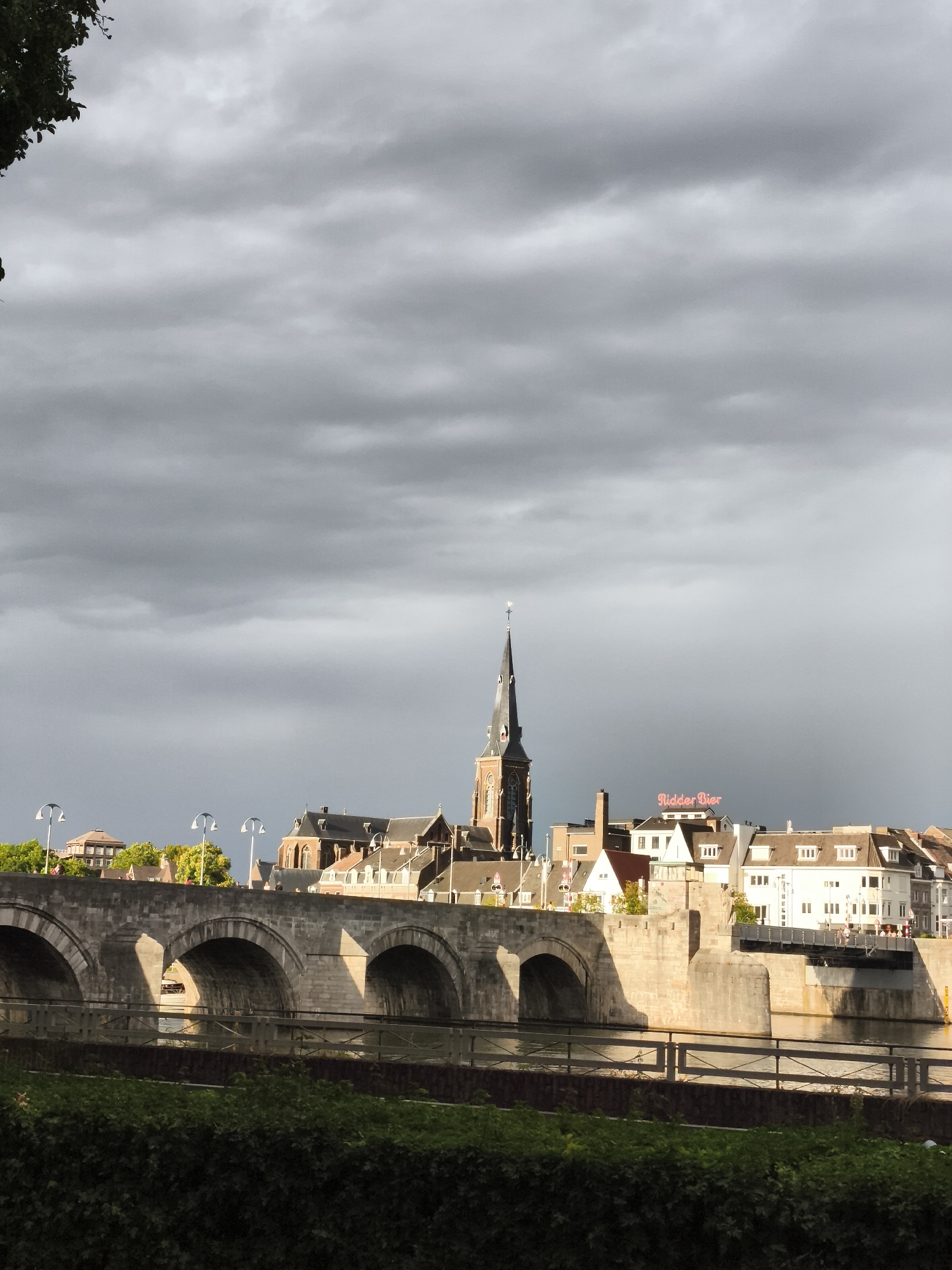 The image shows the Sint Servaasbrug, a historic stone bridge spanning the Maas River in Maastricht, Netherlands. The bridge features multiple arches and is made of stone. On the far side of the river, a cityscape is visible, dominated by a tall church tower with a pointed spire. Several other buildings with tiled roofs and various architectural styles are also present. A neon sign that reads "Ridder Bier" is visible on one of the buildings. The sky is heavily clouded, with dramatic gray clouds suggesting an overcast or approaching storm. In the foreground, a dense green hedge or shrubbery lines the riverbank. Sunlight casts strong shadows on the arches of the bridge, highlighting its structure. A modern pedestrian bridge is also visible to the right of the stone bridge, crossing the river. The scene appears to be captured during the daytime, with the lighting suggesting late afternoon or early evening.