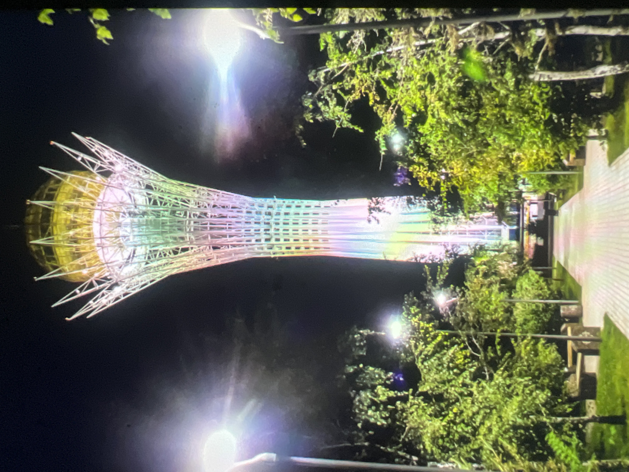 The image depicts the Bayterek Tower in Nur-Sultan, Kazakhstan, at night. The tower, illuminated with rainbow-colored lights, is a prominent structure with a distinctive spherical finial at its apex. It is surrounded by lush green trees and a paved walkway. Several lampposts with bright lights are visible, casting a glow on the scene and contributing to lens flare. The overall ambiance suggests a well-lit urban park or plaza. There are no people visible in the image.