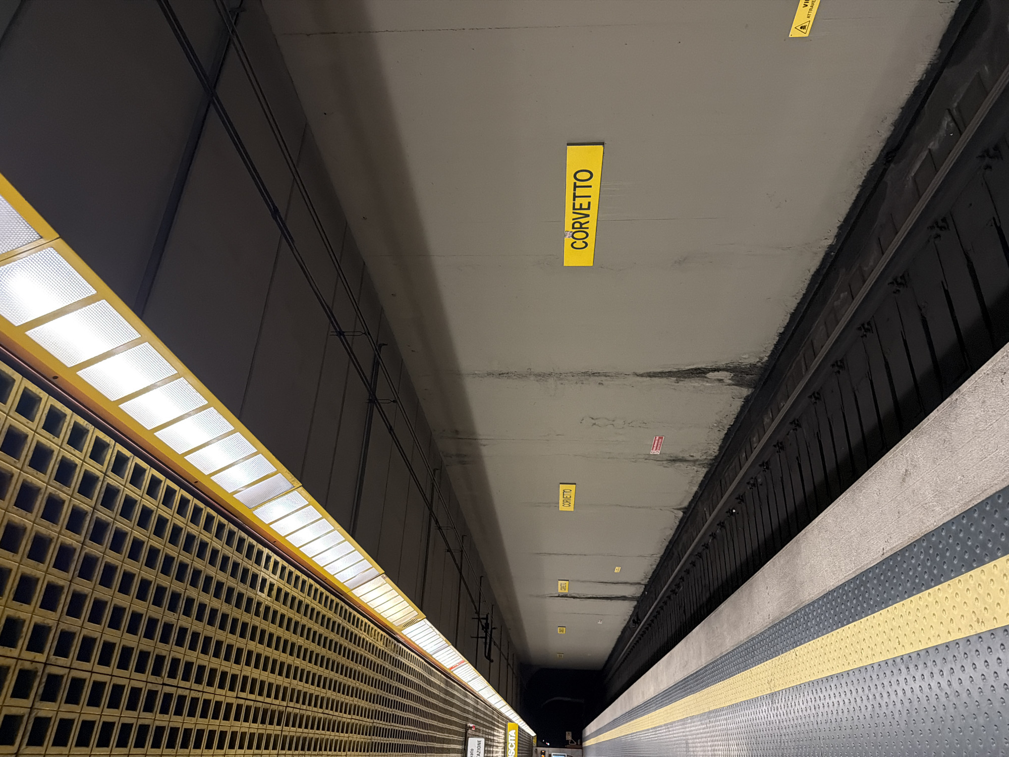 The image depicts a subway station platform in Milan, Italy, specifically the Corvetto station. The perspective is from the platform looking down the tracks towards a tunnel entrance. The platform itself is visible on the left, featuring a textured gray surface with yellow and dark gray guiding lines. Adjacent to the platform are train tracks. The wall of the station is concrete and exhibits some staining. A prominent yellow sign with black text reads "CORVETTO" and is affixed to the wall. Another smaller, partially obscured yellow sign is also visible further down the wall, along with a red warning sign. The ceiling of the station has a distinctive grid-like structure made of yellow material, with illuminated panels interspersed. Overhead, electrical wires for the train system are visible. The overall atmosphere is utilitarian and characteristic of a public transportation hub.