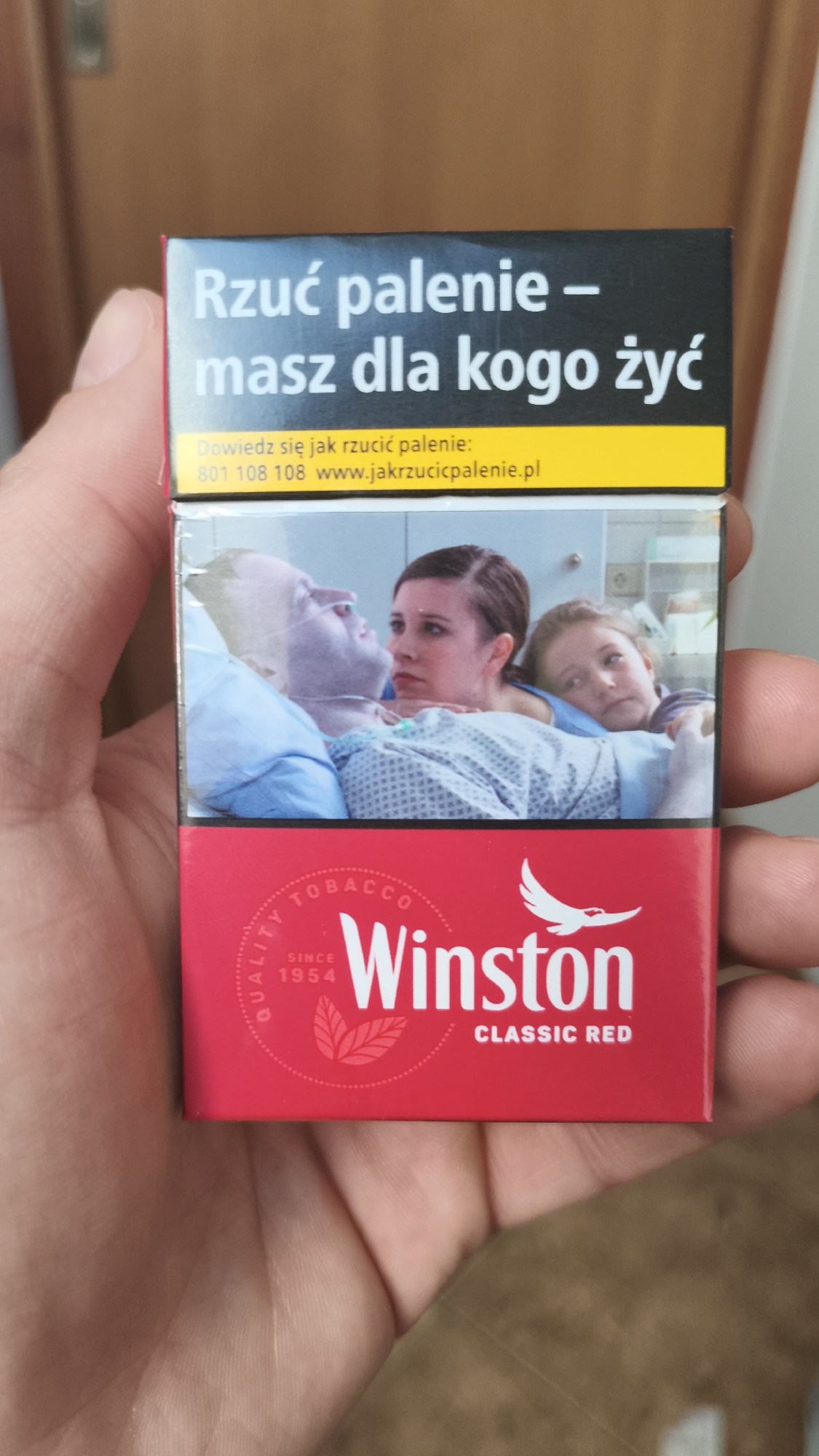 A hand holds a pack of Winston Classic Red cigarettes. The top of the pack has a black box with yellow text that reads, "Rzuć palenie - masz dla kogo żyć" with information about how to quit smoking below. Below that is an image of a sick person in a hospital bed, with a woman and child next to them. The Winston logo is at the bottom. There is no explicit information indicating the image was taken in Nowy Tomyśl, Poland.