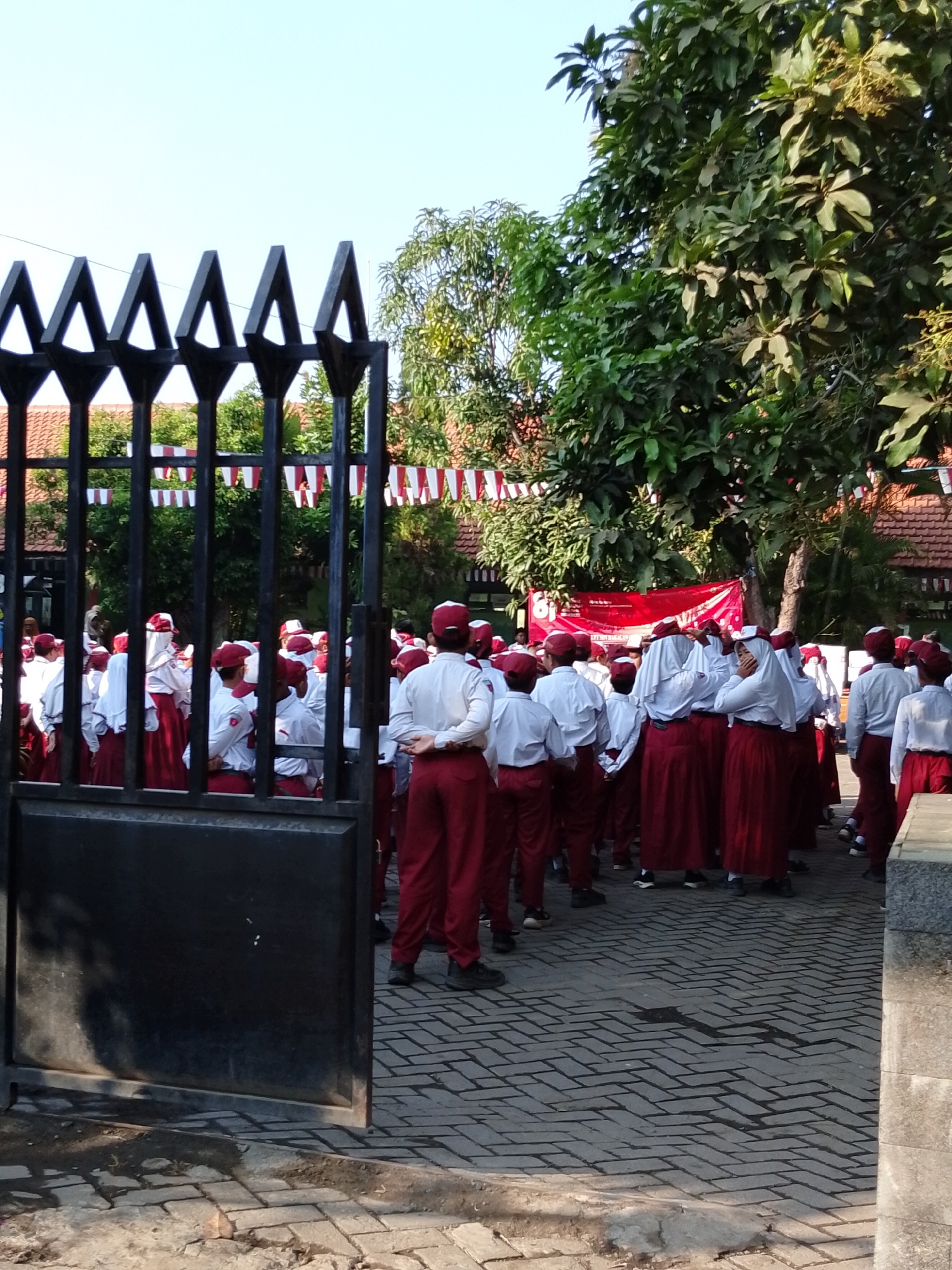 The image shows a group of Indonesian schoolchildren, likely elementary school age, gathered in an outdoor area. They are wearing white shirts with maroon pants and skirts, and many have maroon caps. Some of the children are wearing white headscarves over their caps. They appear to be standing in rows, facing forward, possibly for a school assembly or flag ceremony, as indicated by the red and white banner in the background, which is common during Indonesian Independence Day celebrations. The setting appears to be a schoolyard, with a paved walkway made of interlocking bricks, trees, and buildings with red tiled roofs in the background. A black metal gate is in the foreground, partially obscuring the view of the students. The sky is clear and blue, suggesting it is a sunny day. The scene is likely located in the city of Pasuruan, Indonesia, as indicated by the context.