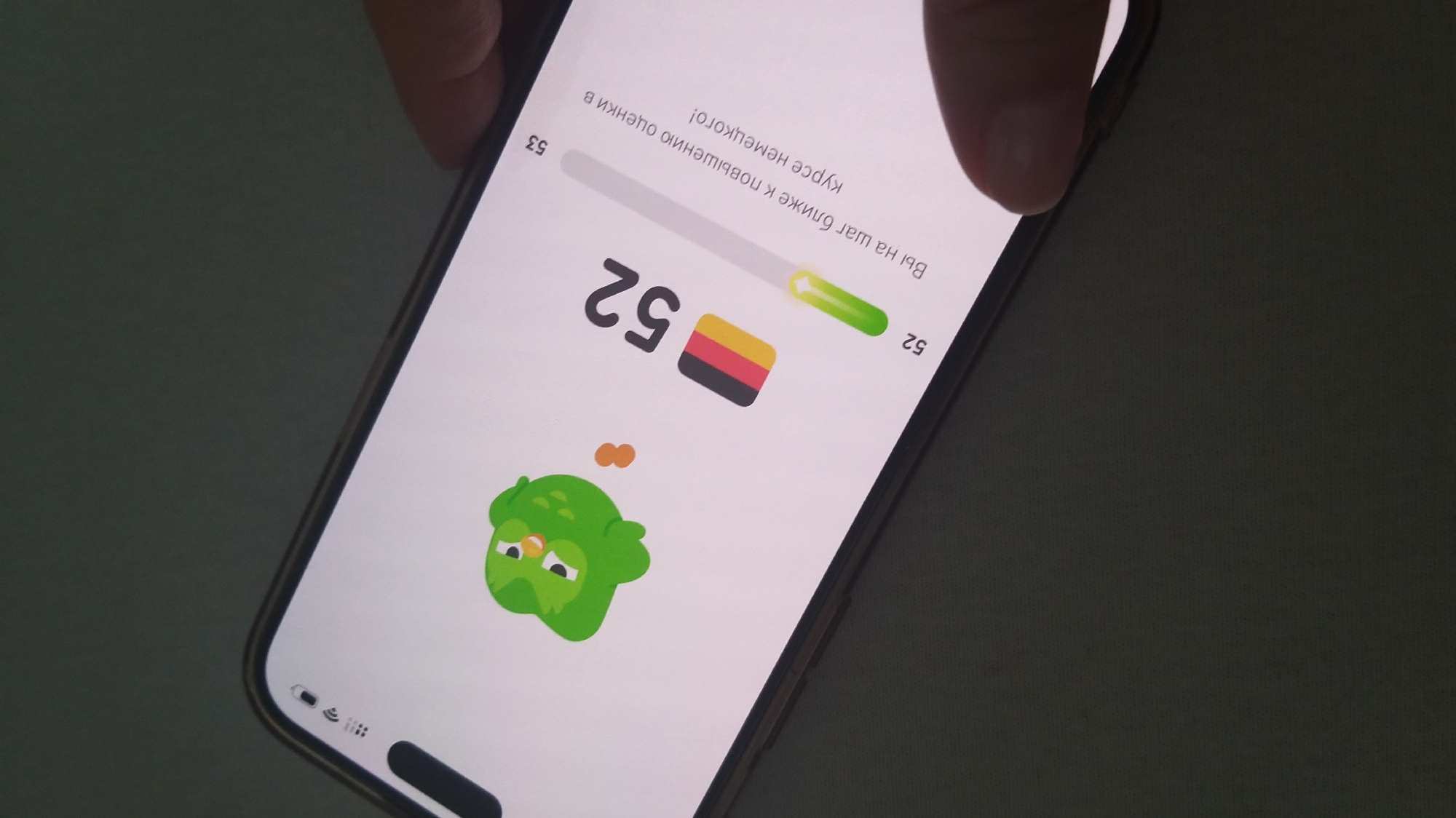 A phone screen displays the Duolingo app, showing a green owl character and progress towards level 53 in German language. The current level is 52, indicated by a progress bar. The text on the screen reads "Вы на шаг ближе к повышению оценки в курсе немецкого!", which translates to "You are one step closer to improving your grade in the German course!". The phone is held by a hand, and a person's thumb is visible on the right side of the screen. The background is dark and out of focus, suggesting the image was taken indoors. There is no information in the image that indicates a specific location in Krefeld, Germany.