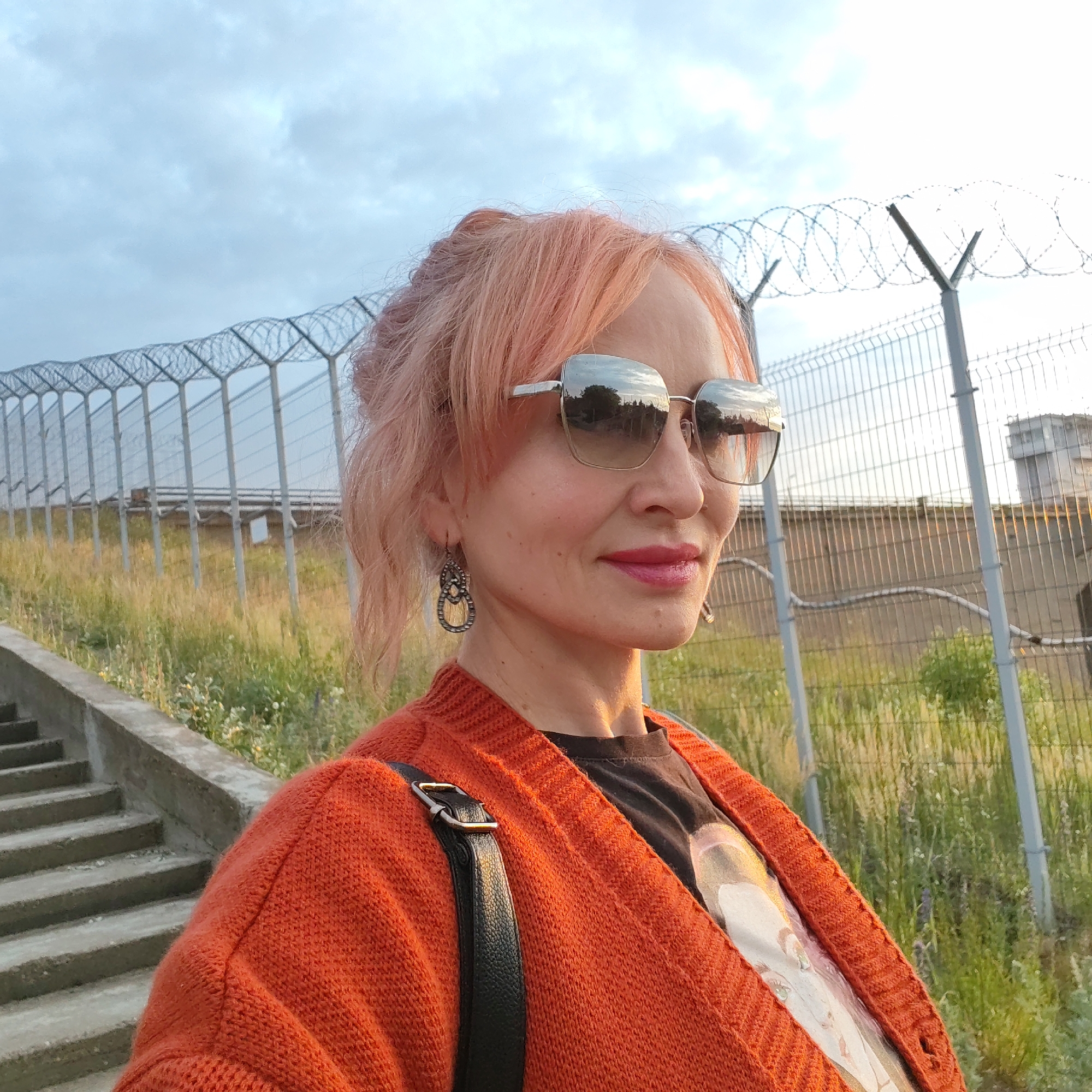 A medium shot, taken outdoors at dusk, features a woman with light pink hair and sunglasses, looking to her right with a slight smile. She is wearing an orange knitted cardigan over a dark t-shirt. A black strap from a bag crosses her chest. The background includes a chain-link fence with barbed wire on top, a grassy slope, and a concrete staircase. In the distance, a building with a tower is visible. The sky is overcast with clouds. This appears to be a public area, possibly near a facility with security fencing, within the city of Chaikovsky, Russia.