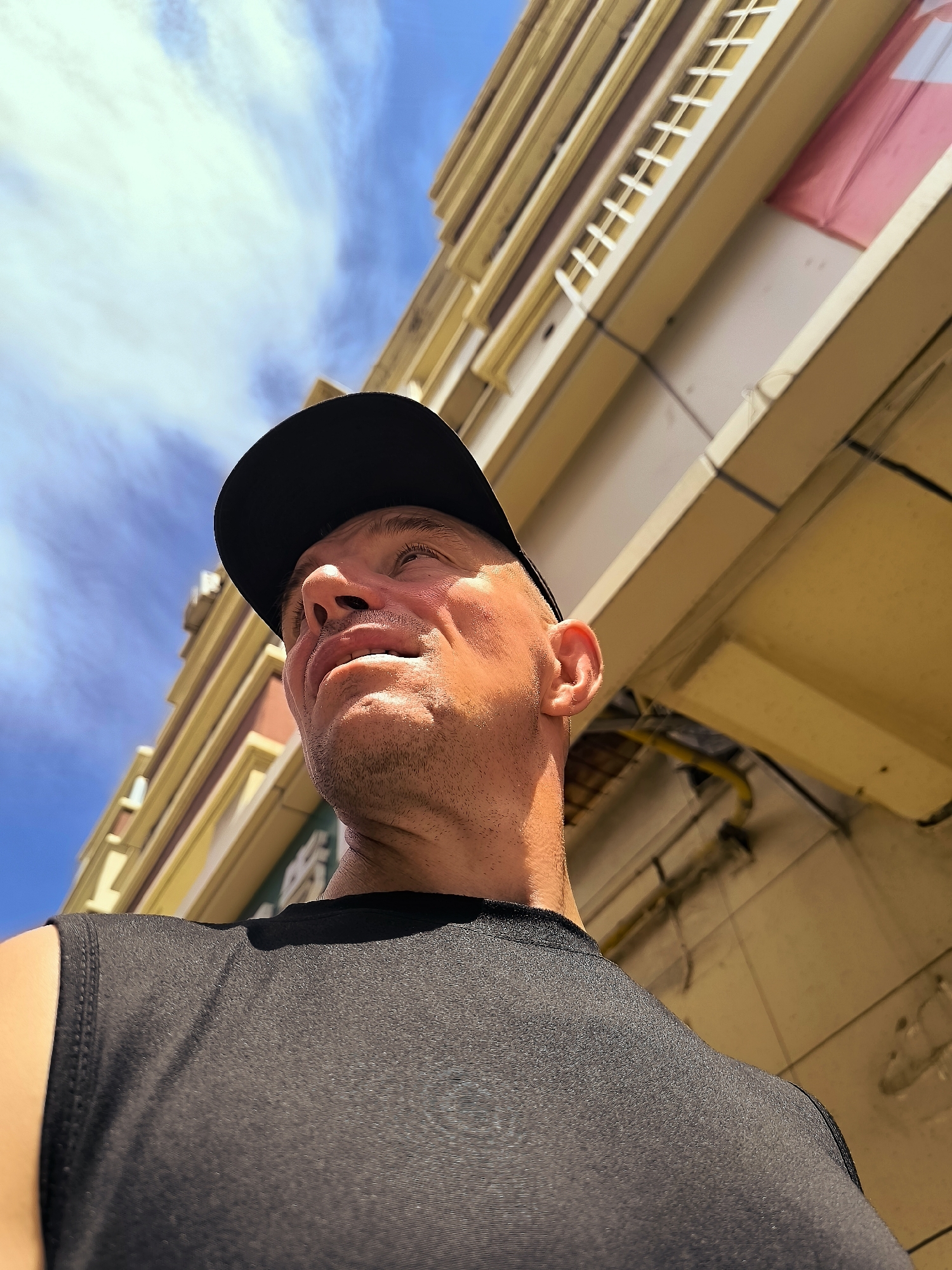 An upward-angled, close-up shot captures a fair-skinned man wearing a black baseball cap and a black sleeveless shirt. He is looking upwards and to his left, with his mouth slightly open. His face is illuminated by sunlight, highlighting his features and the stubble on his chin. Behind him, a cream-colored building with yellow trim and a pink facade is visible against a bright blue sky with wispy clouds. The building's architecture suggests it is likely in Ürümqi, China, given the style of the window frames and decorative elements. The man appears to be outdoors in an urban setting.