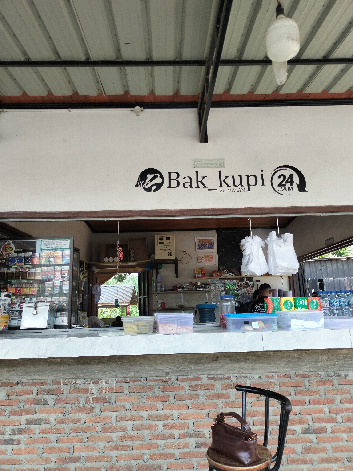 The image depicts the exterior of a small shop, likely a roadside stall or convenience store, with a counter and merchandise visible. The shop's signage reads "Bak_kupi 24 JAM -EH MALAM-", suggesting it sells coffee and operates 24 hours, possibly specializing in nighttime service. The building has a corrugated metal roof supported by dark metal beams. Behind the counter, an open doorway reveals a glimpse of the outside, with greenery and a roof structure of another building.

On the counter, various items are displayed, including bottles of water, packaged snacks, and plastic containers. To the left, a glass-fronted refrigerator is filled with numerous cigarette packs. A person, likely an employee, is partially visible behind the counter, attending to customers or restocking. In the foreground, a dark brown leather handbag rests on a wooden stool with a black metal frame. The overall impression is of a modest, functional establishment serving local needs in a semi-rural or suburban setting. The wall in the foreground is constructed of red brick. The location is identified as Kutalawah, Indonesia.