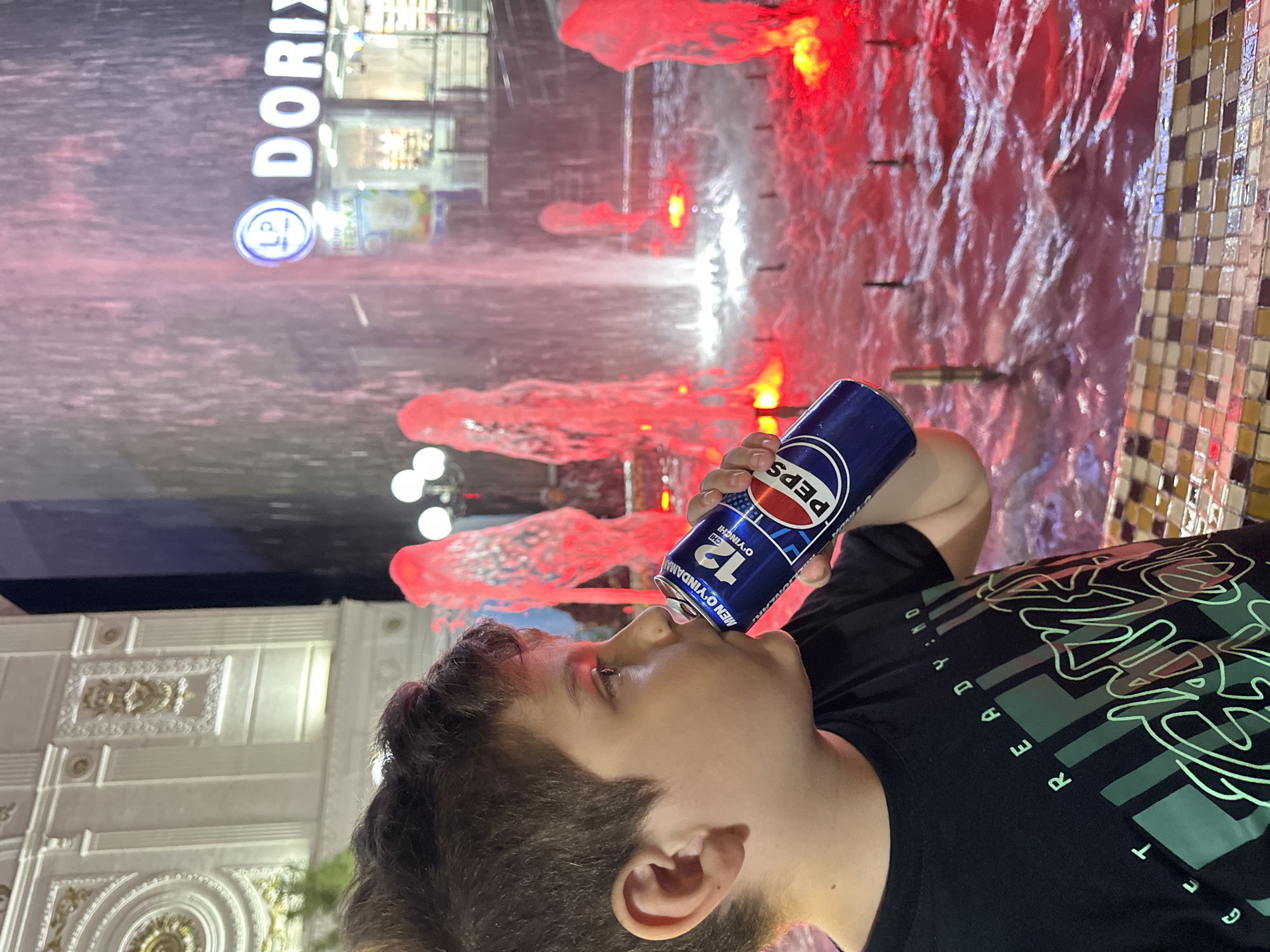 A boy is drinking a can of Pepsi in front of a fountain with red lights in Tashkent, Uzbekistan. The boy is wearing a black t-shirt with a graphic design. In the background, there is a building with ornate architecture and a storefront with the logo "DORIX" visible. The fountain water is illuminated by red lights, creating a vibrant and dynamic scene. The image appears to be taken at night.