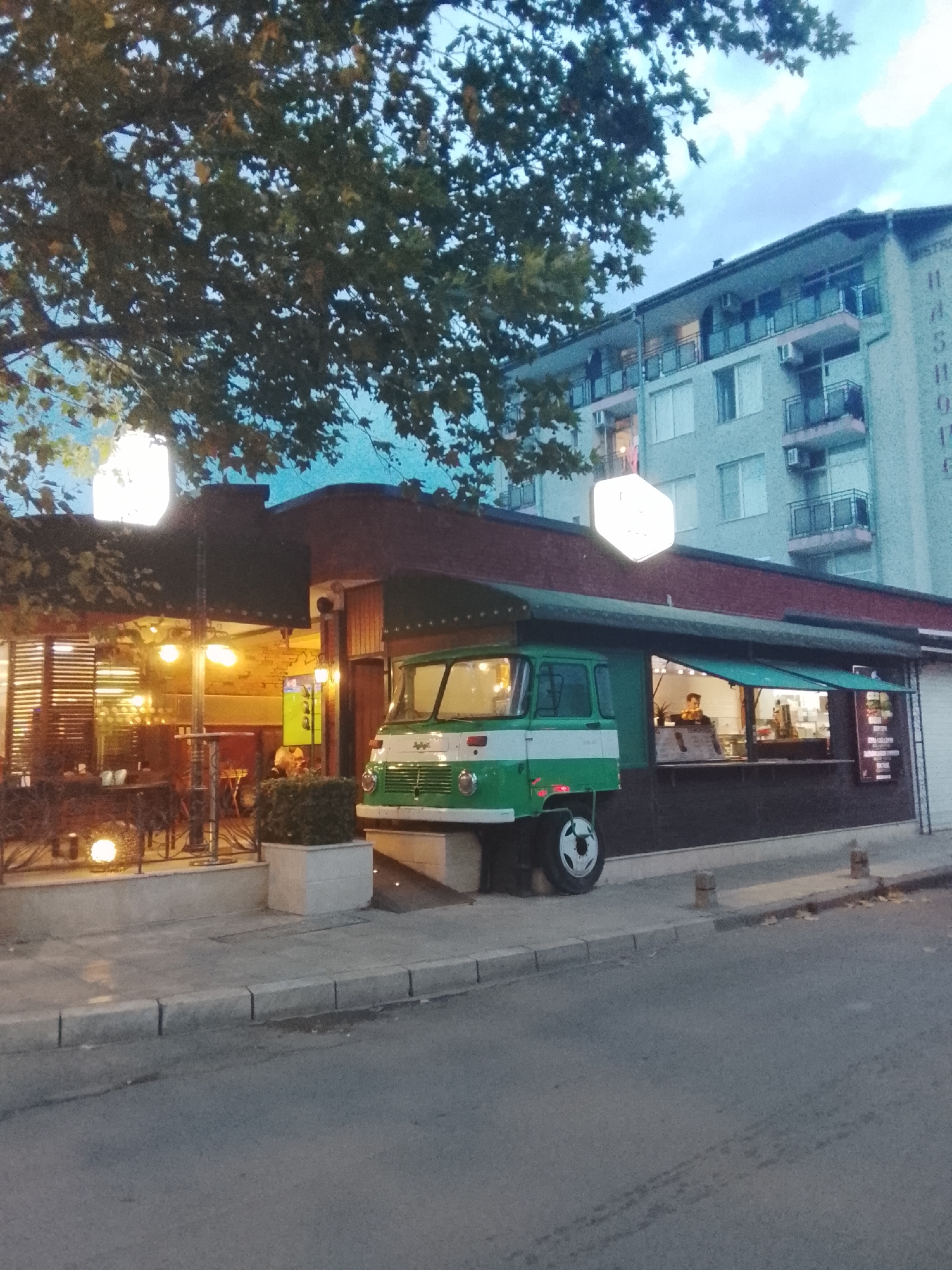 The image shows a street scene in Nesebar, Bulgaria, during what appears to be twilight. In the foreground, a gray asphalt road with white lane markings is visible, bordered by a concrete sidewalk with curbs. A portion of a green and white truck, possibly a food truck, is parked on the sidewalk. The truck has a retro design, with large circular headlights and a prominent grille.
Behind the truck, a low-rise building with a dark red roof and brown siding extends along the street. This building appears to house a business with an outdoor seating area, indicated by tables and chairs visible under a covered section. Several illuminated globes are placed around this area, casting a warm light. Further back, a multi-story apartment building with a light blue facade rises above the lower structure. The building has numerous windows and balconies.
A large tree with dark green foliage partially obscures the sky and the upper portion of the apartment building. The sky is a dusky blue, with some clouds visible. The overall impression is a residential or commercial street in an urban environment, with a distinct architectural style suggesting a European location. The lighting suggests it is either early morning or late evening.