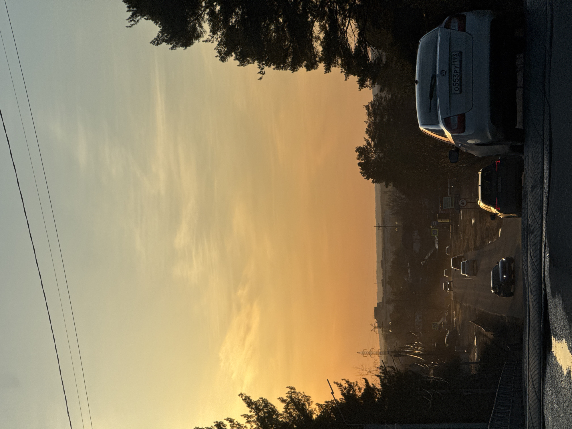 This is an outdoor shot of a cityscape. The sky, ranging from light blue to orange, indicates either sunrise or sunset. Power lines run through the top left of the shot. Trees frame the sky, providing a natural border at the top and bottom.
On the right side of the image, cars are parked next to a road. Traffic is visible moving along the road, heading into the city. License plate: 0553РУ 193.
The location is the city of Chaikovsky, Russia.