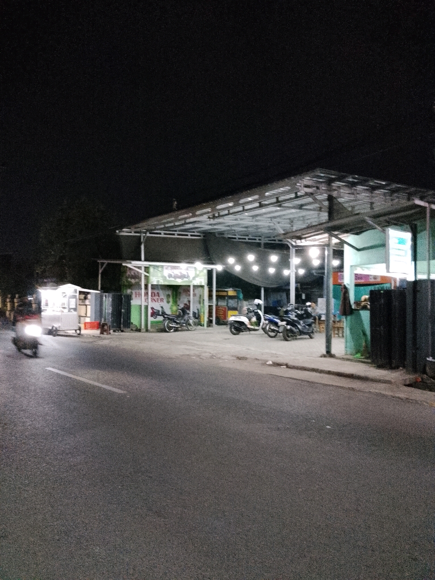 The image captures a nighttime scene on a street in Pasuruan, Indonesia, likely in front of a food stall or small eatery. A brightly lit food cart is visible on the left, with a motorcycle passing by, its headlights illuminated. To the right, a covered structure with multiple hanging lights illuminates a street-side establishment. Several motorcycles are parked under this structure, suggesting it's a common gathering spot. The area appears to be a commercial or semi-commercial zone, typical of roadside businesses in Indonesian cities. The overall impression is one of a lively, local nighttime commercial area.