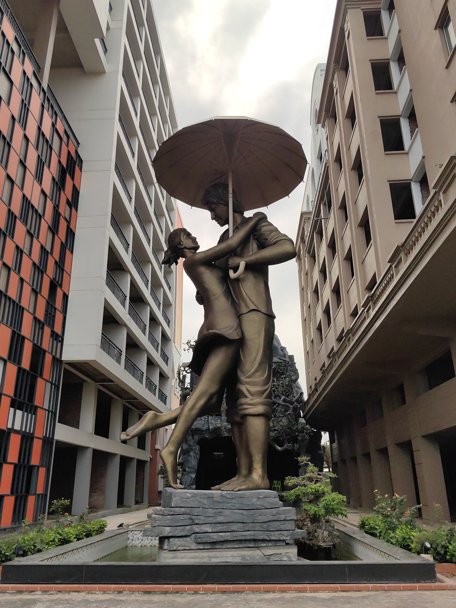The image shows a bronze statue of a couple embracing under an umbrella, located in Tuan Chau Ward, Vietnam. The male figure is standing, while the female is leaning into him with one leg raised. They stand on a stone platform surrounded by greenery and a small water feature. The statue is positioned between buildings, one with a modern orange and black facade, another a light brown building with classical architectural details, and a white building. The sky above is overcast.