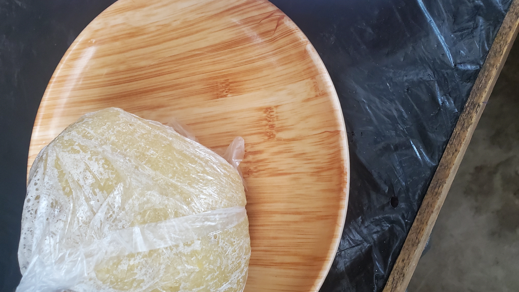 A round, light-yellow food item, possibly dough or a starchy preparation, is sealed in a clear plastic bag and rests on a wooden plate. The plate has a natural wood grain pattern with vertical lines and a light brown hue. The plate is positioned on a surface covered by black plastic sheeting. The scene appears to be outdoors, with a portion of a wooden plank visible at the bottom edge of the frame. The lighting suggests it is daytime. No specific landmarks or urban features are visible to pinpoint a location within Jalingo, Nigeria.