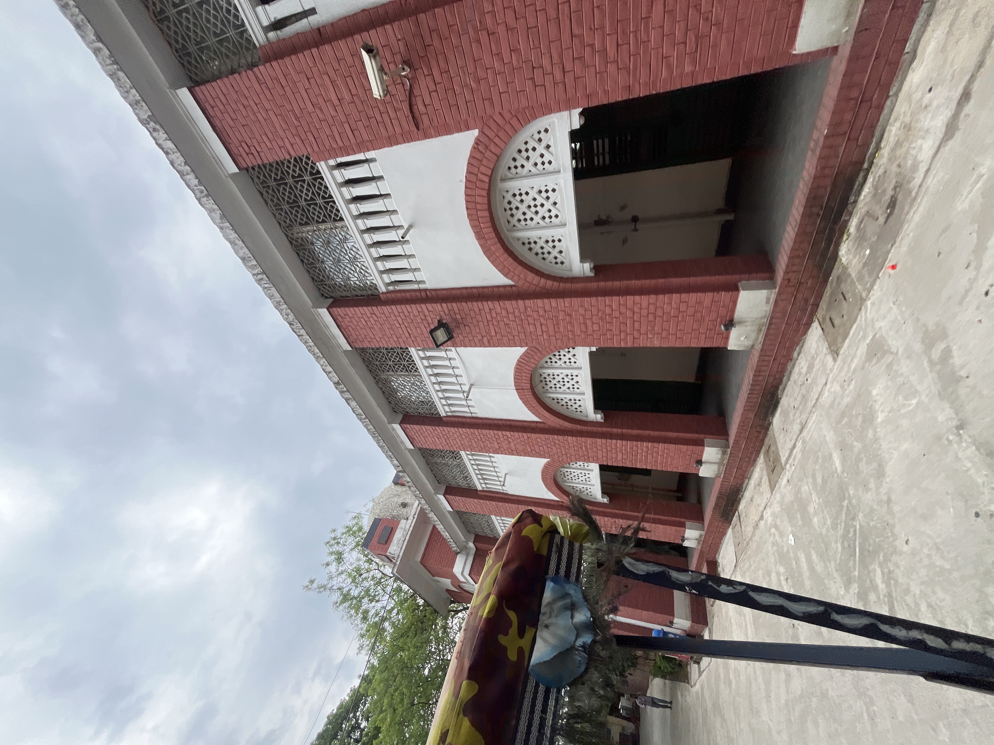 The photograph shows a brick building in Dhaka, Bangladesh. The building is two stories high, with a red brick facade and white trim. There are arched openings on the second story, and the windows have decorative ironwork. A surveillance camera is mounted near the top right. In the foreground, there is a colorful banner or decoration of some kind. The sky is overcast. The photo is taken from a low angle, looking up at the building.