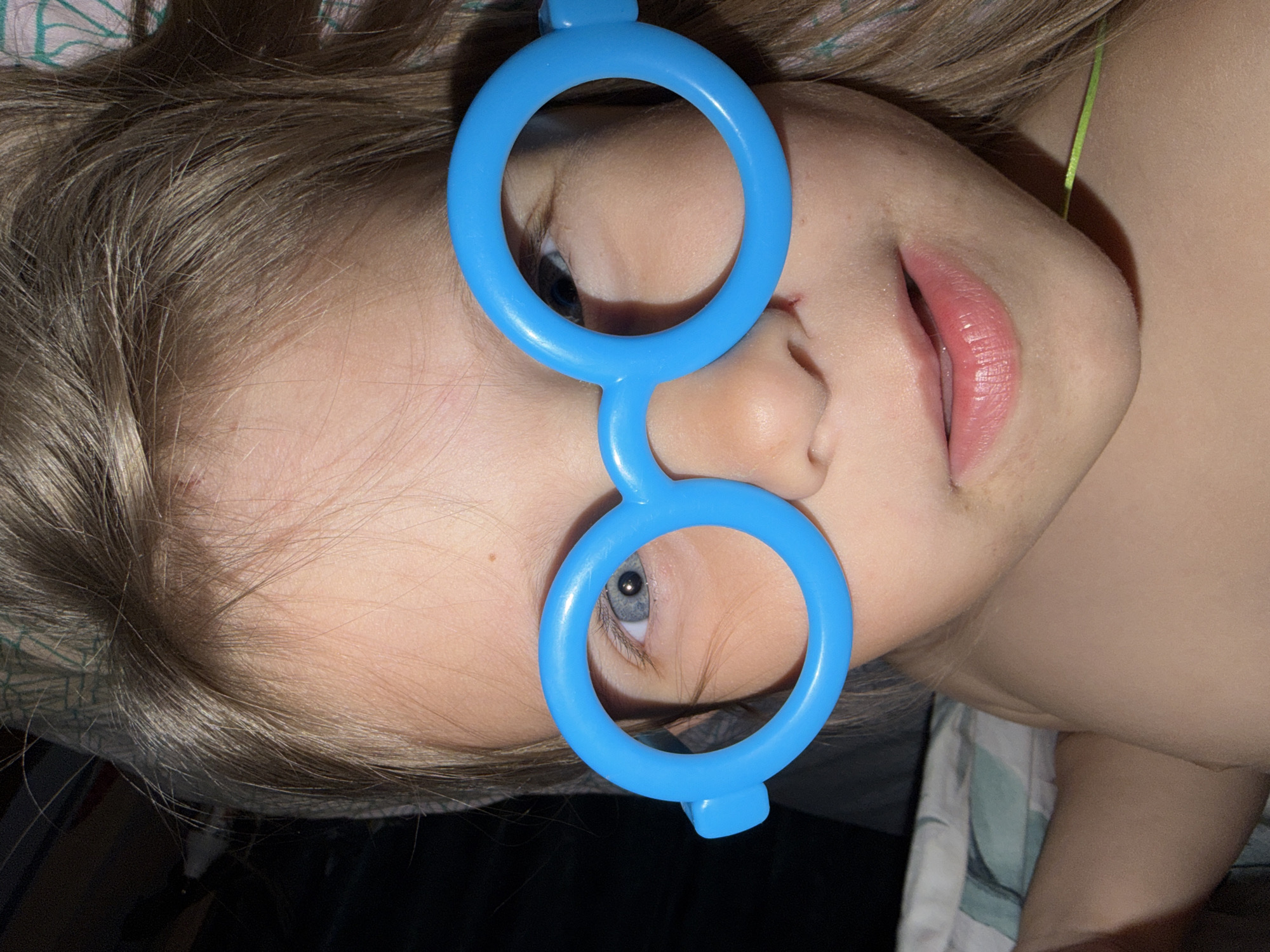A close-up, rotated portrait captures a young child lying down, looking directly towards the viewer. The child has fair skin and light brown hair, which frames their face. They are wearing oversized, bright blue toy glasses with large, round frames that cover a significant portion of their face, framing their blue-gray eyes. Their lips are slightly parted, revealing a light pink color. A thin, bright green string or ribbon is visible near the child's right ear. In the background, elements of patterned fabric, possibly bedding or clothing, are visible, suggesting an indoor, home setting in Chaikovsky, Russia. The image has neutral indoor lighting. The child's expression is calm and observant, conveying a sense of playful innocence. No text is visible in the image.