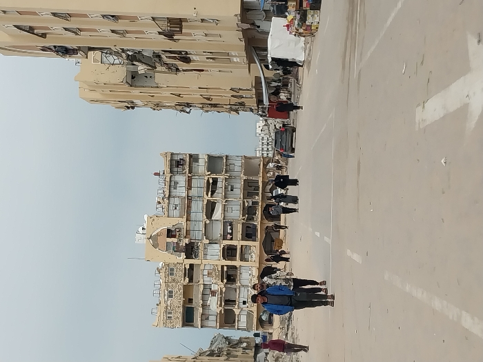 A wide street divides damaged buildings, evidence of conflict. The structures are heavily scarred, with walls missing and interiors exposed. People walk along the street, navigating the aftermath. Vehicles are parked further up the road. A makeshift white structure contrasts with the damaged surroundings, possibly providing temporary shelter. The scene conveys destruction and resilience amid the backdrop of urban devastation.