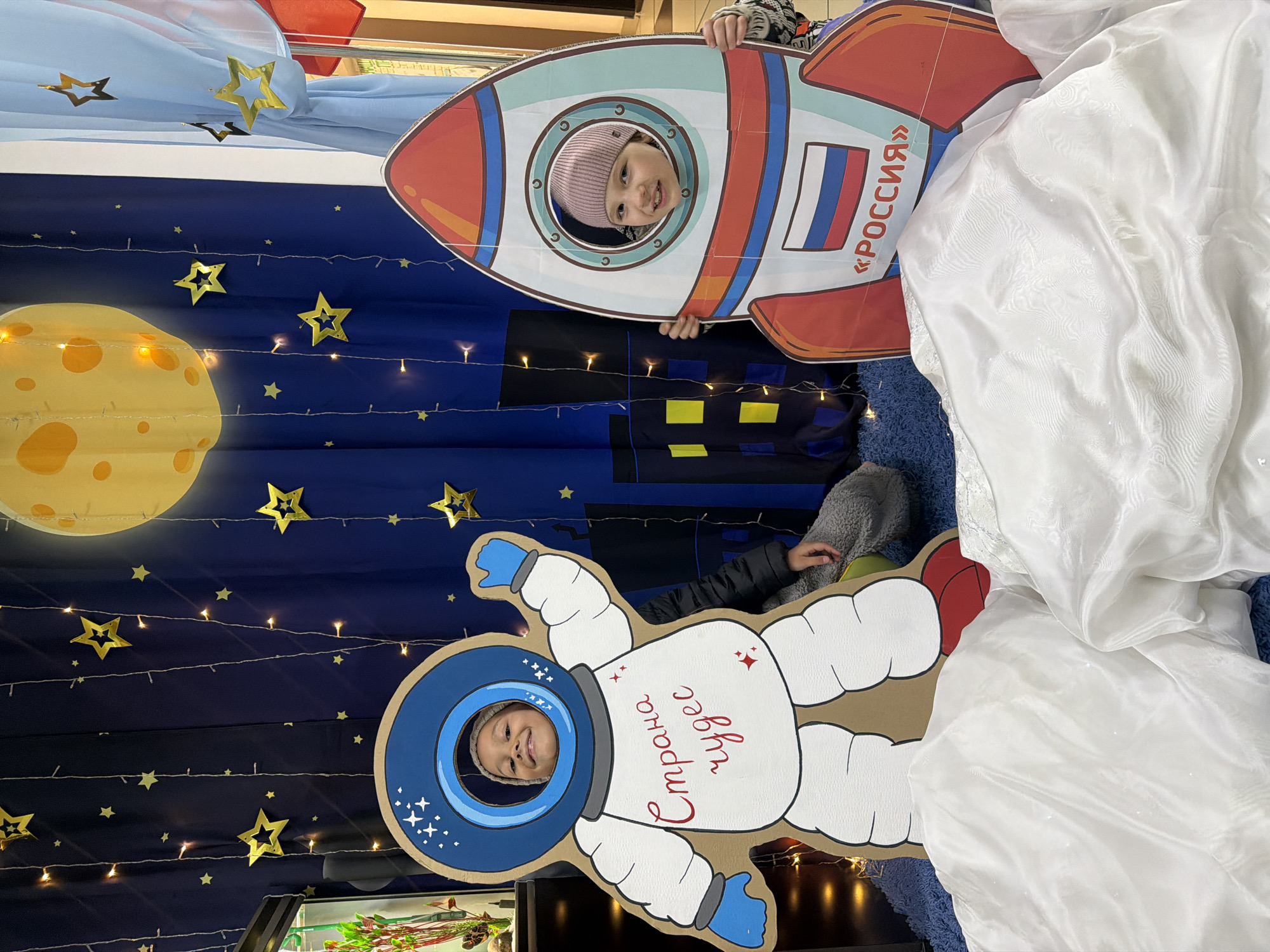 Two children are posing with cardboard cutouts related to space exploration. A girl wearing a pink hat is positioned behind a rocket cutout that features the colors of the Russian flag and the word "Россия" (Russia). Below her, another child is behind an astronaut cutout, with the Russian text "Страна Чудес" (Wonderland) written on the body. The backdrop behind them includes a dark blue cloth adorned with golden stars and a large, illuminated moon. The scene is also decorated with string lights. In the background, there are hints of an indoor setting, possibly a store or event space. According to the metadata, the location is in the city of Krasnoyarsk, Russia.