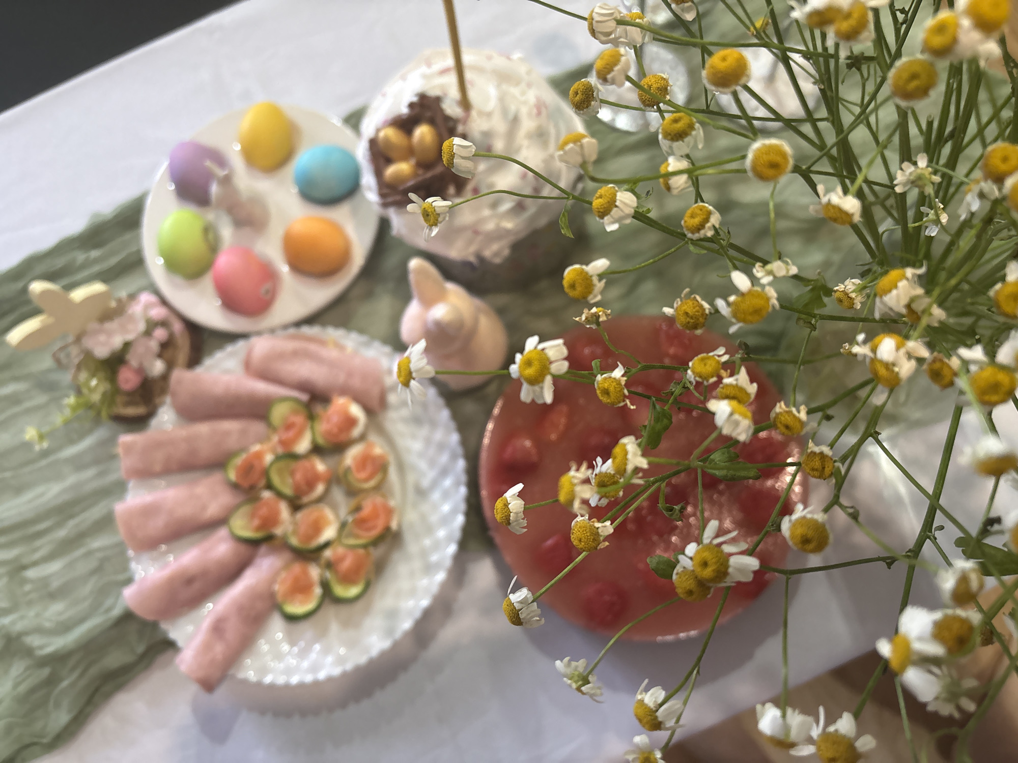 The scene depicts an Easter-themed table arrangement. There is a white plate holding multiple colored Easter eggs, a glazed Easter cake containing a nest with chocolate eggs, a plate of sliced ham alongside canapés of cucumber topped with salmon, a clear red jelly dessert with embedded fruit, a ceramic bunny figurine, a floral arrangement with small daisies, and a green wrinkled cloth. The location is not discernible as being specifically in Chaikovsky, Russia.